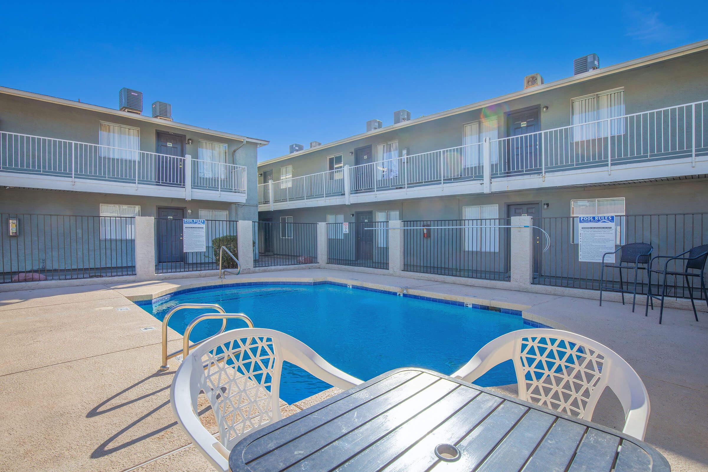 A well-maintained outdoor swimming pool surrounded by a courtyard with two-story buildings. Lounge chairs and a table are set up near the pool. The clear blue sky reflects a sunny day, enhancing the inviting atmosphere of the area.