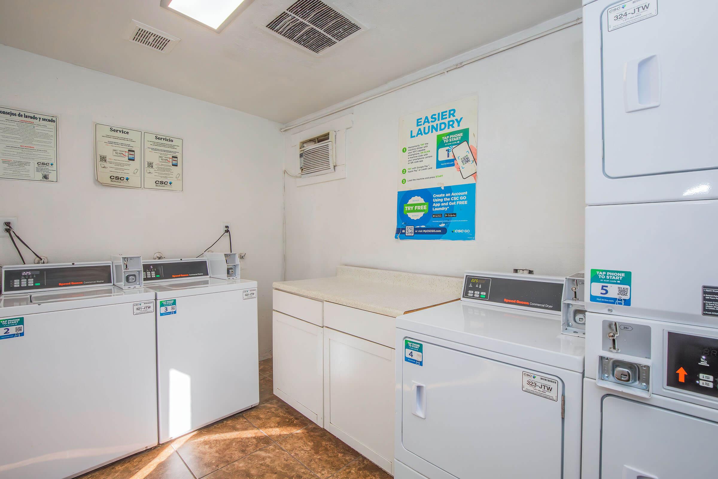 A clean laundry room featuring several white washing machines and dryers. There is a countertop for folding clothes, along with informational posters on the wall about laundry services. The floor is tiled, and the space is well-lit with a ceiling vent for ventilation.