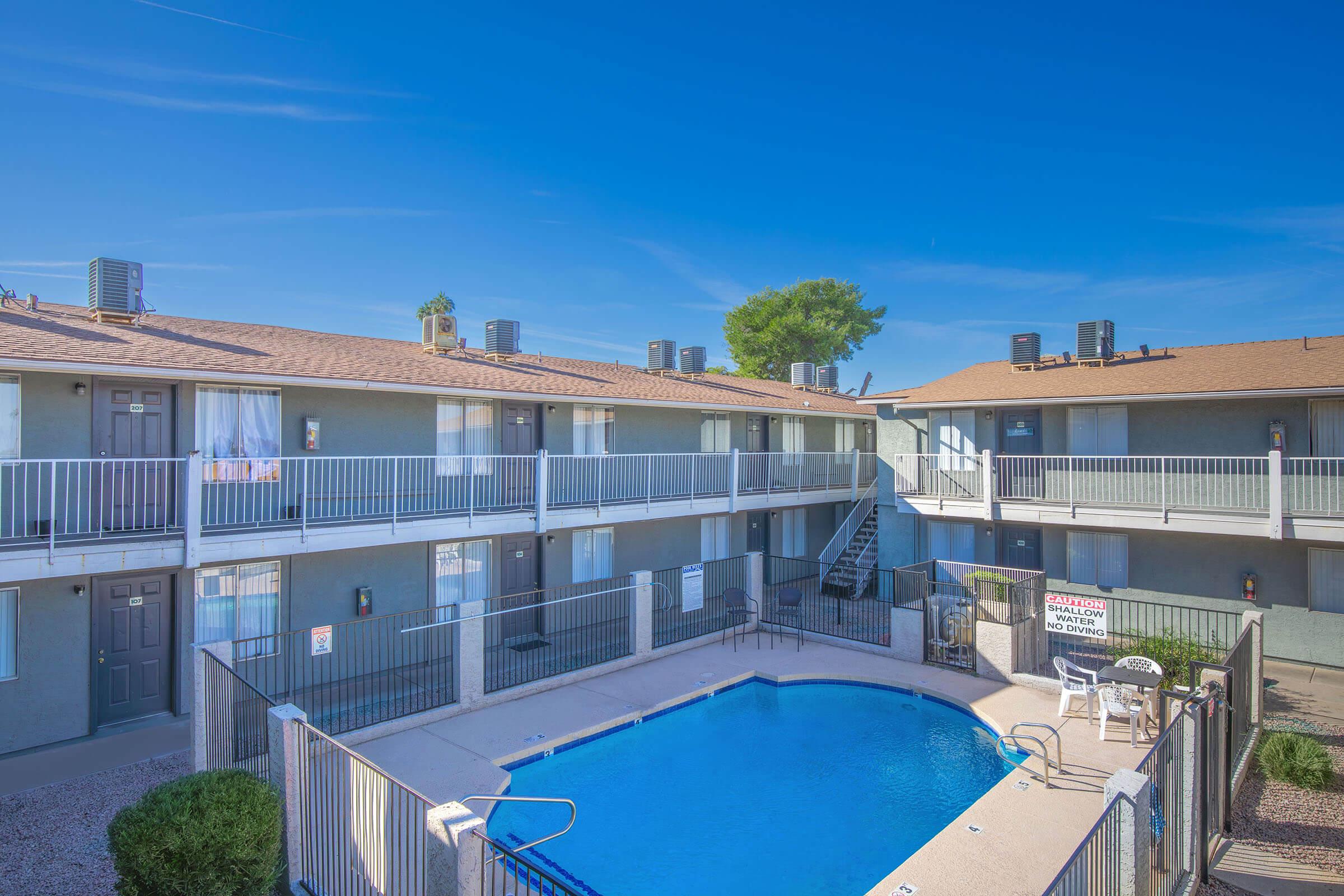 A view of a courtyard featuring a swimming pool surrounded by a two-story apartment building with balconies. The sky is clear and blue, and there are several air conditioning units visible on the roofs. The area is well-maintained with landscaping around the poolside.