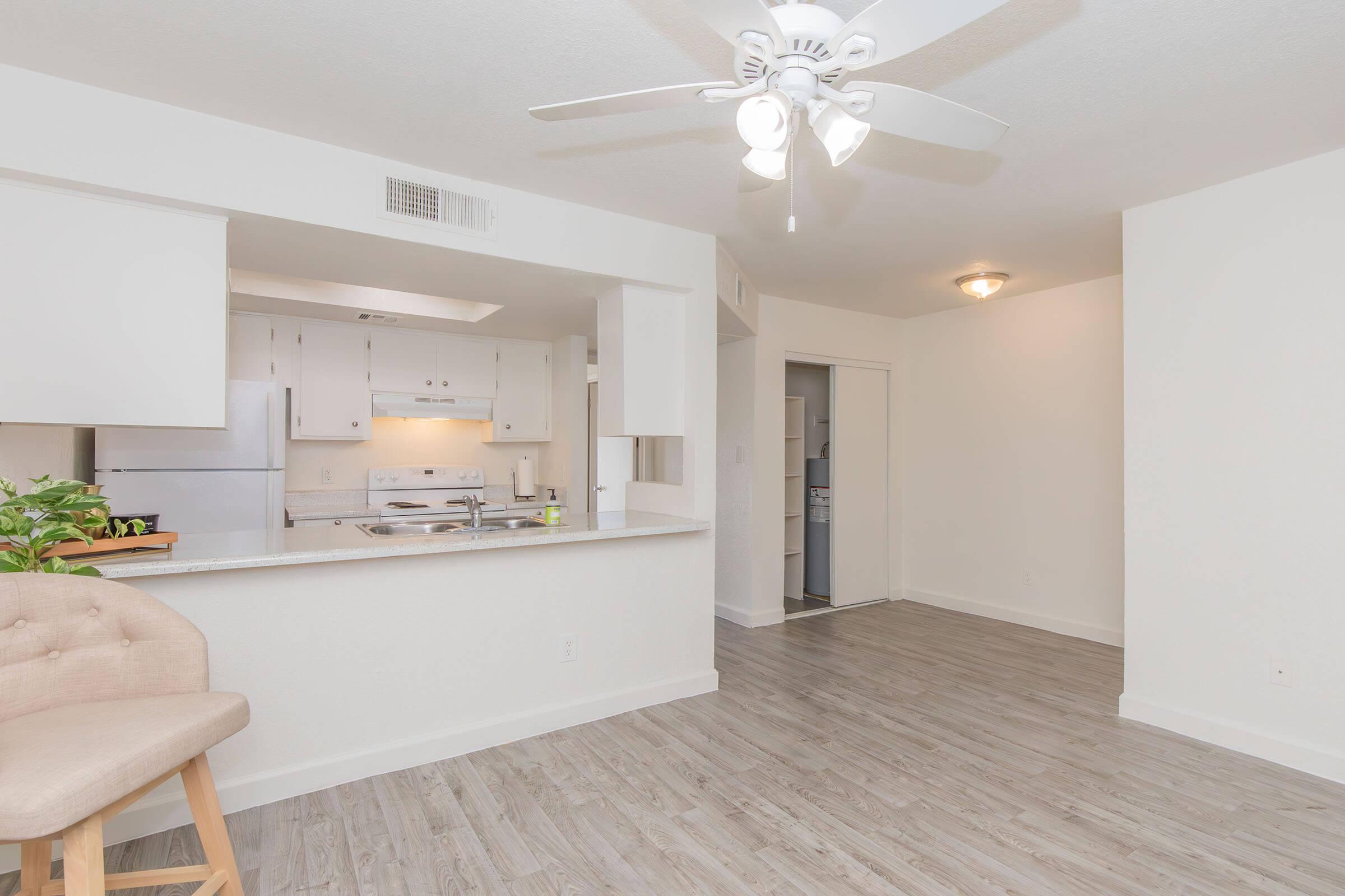 A spacious, modern apartment interior featuring an open layout. The kitchen area is visible with white cabinets and appliances, and a small bar counter with a beige stool. A ceiling fan is overhead, and the light-colored flooring complements the bright walls. The living space is empty and well-lit.