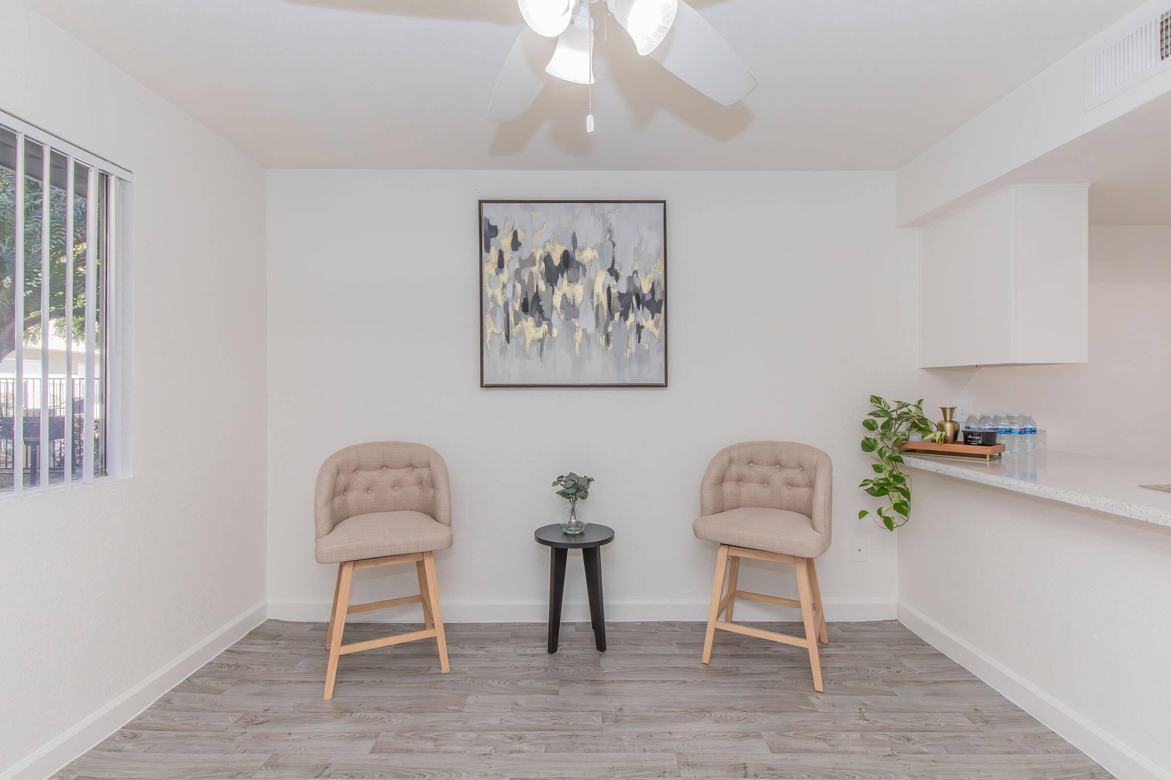 Two beige upholstered chairs are positioned on a light wooden floor, facing a small black side table with a single green plant. On the wall hangs an abstract painting in shades of gray, yellow, and black. A white ceiling fan is visible, and a window with vertical blinds allows natural light into the room.