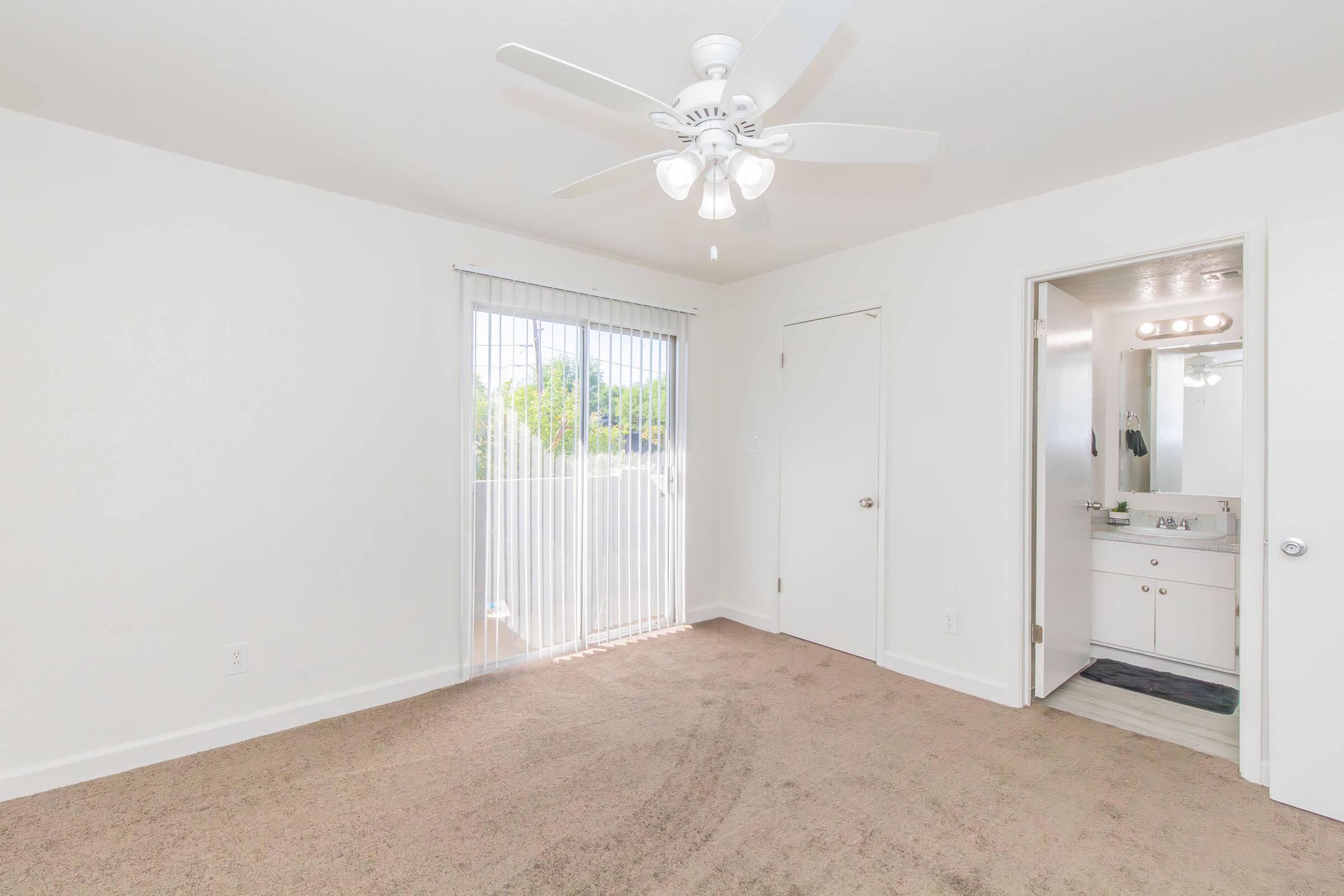 A well-lit, empty bedroom with beige carpet, a ceiling fan, and a sliding glass door leading to a balcony. A bathroom door is visible to the right, featuring a single vanity and a mirror with lights above. The walls are painted white, creating a bright and airy atmosphere.