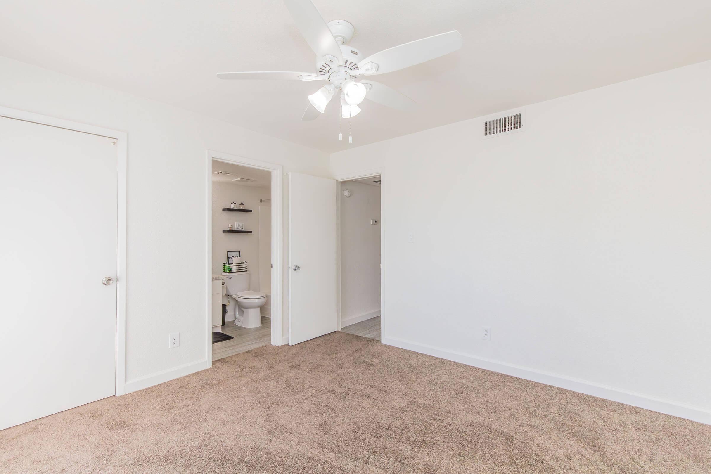 A bright, empty room with light beige carpet and white walls. A ceiling fan is visible above. There are two doorways: one leading to a bathroom with a toilet visible in the background and the other to a different space. Natural light illuminates the room, creating an inviting atmosphere.
