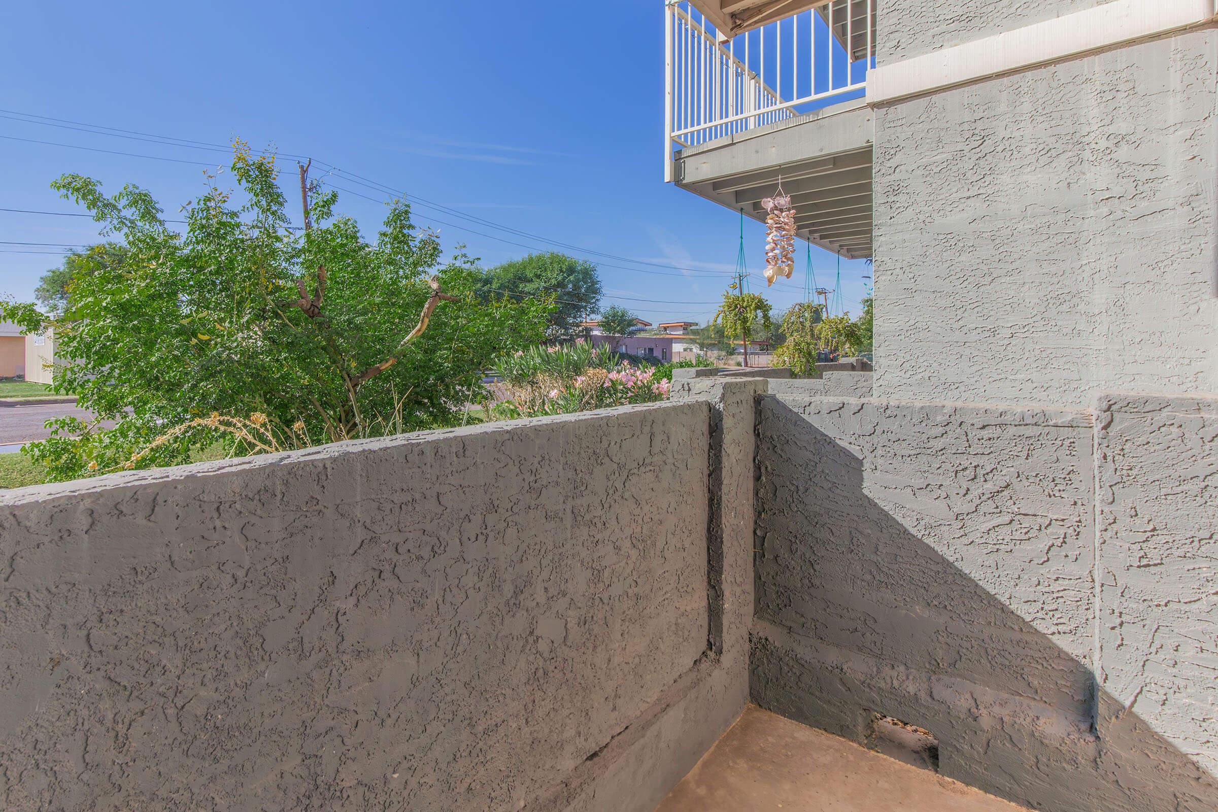 A view from a balcony overlooking a quiet street, with green trees and bushes visible. The surrounding area is sunny, and a portion of a neighboring building and the sky are also visible. The wall texture is rough, indicating a simple concrete structure.