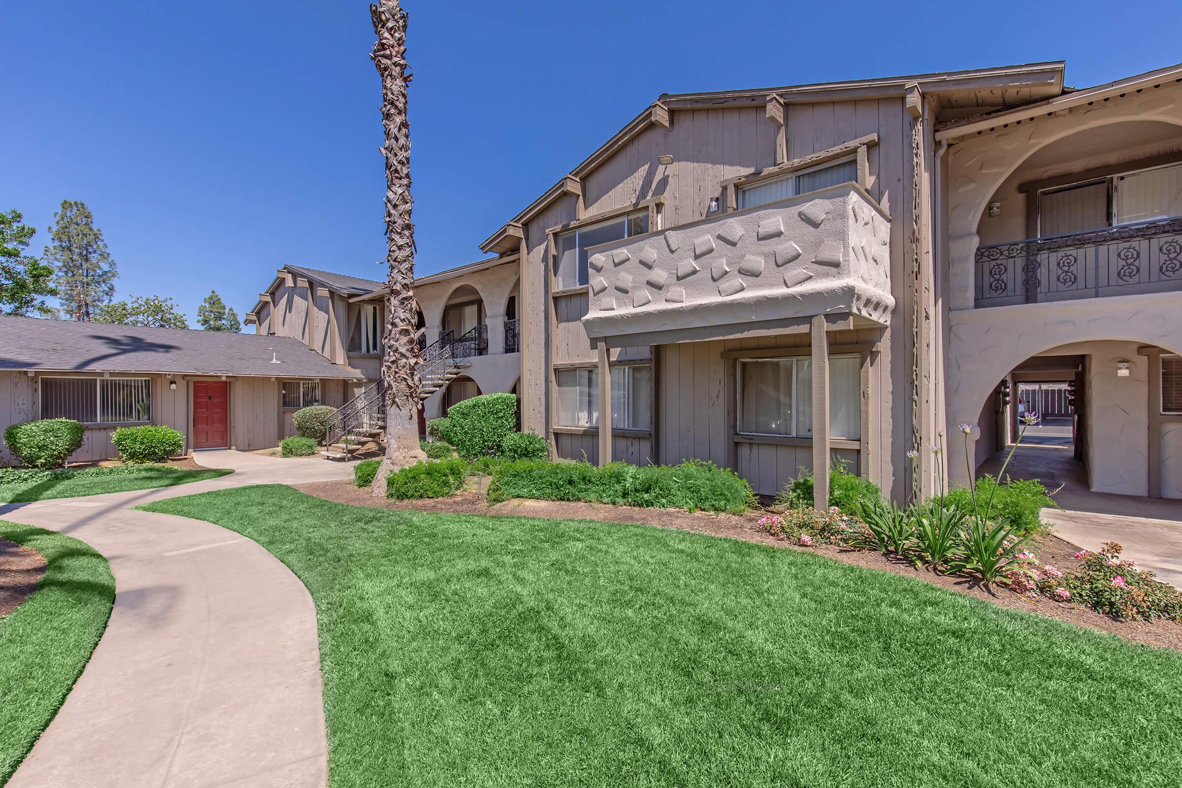 A well-maintained apartment complex featuring modern architecture, surrounded by lush green lawns and neatly trimmed bushes. The pathway curves through the landscaped area, leading to the entrances of various units under a clear blue sky. Palm trees add a touch of tropical ambiance to the scene.