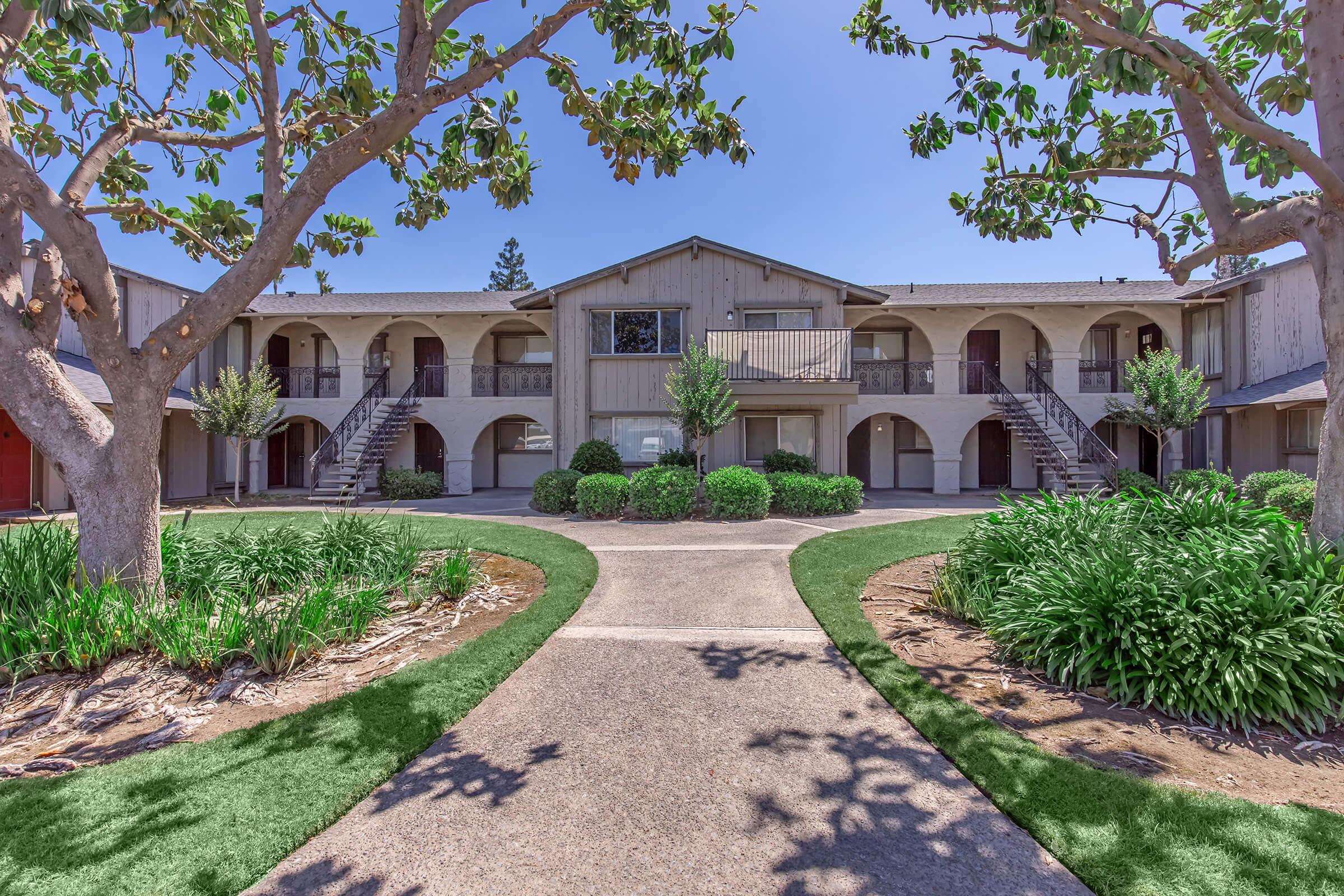 A view of a two-story apartment complex surrounded by lush greenery and landscaped paths. The building features multiple entrances with staircases on either side, and small trees and shrubs enhance the outdoor aesthetic. The scene is bright and inviting under clear blue skies.