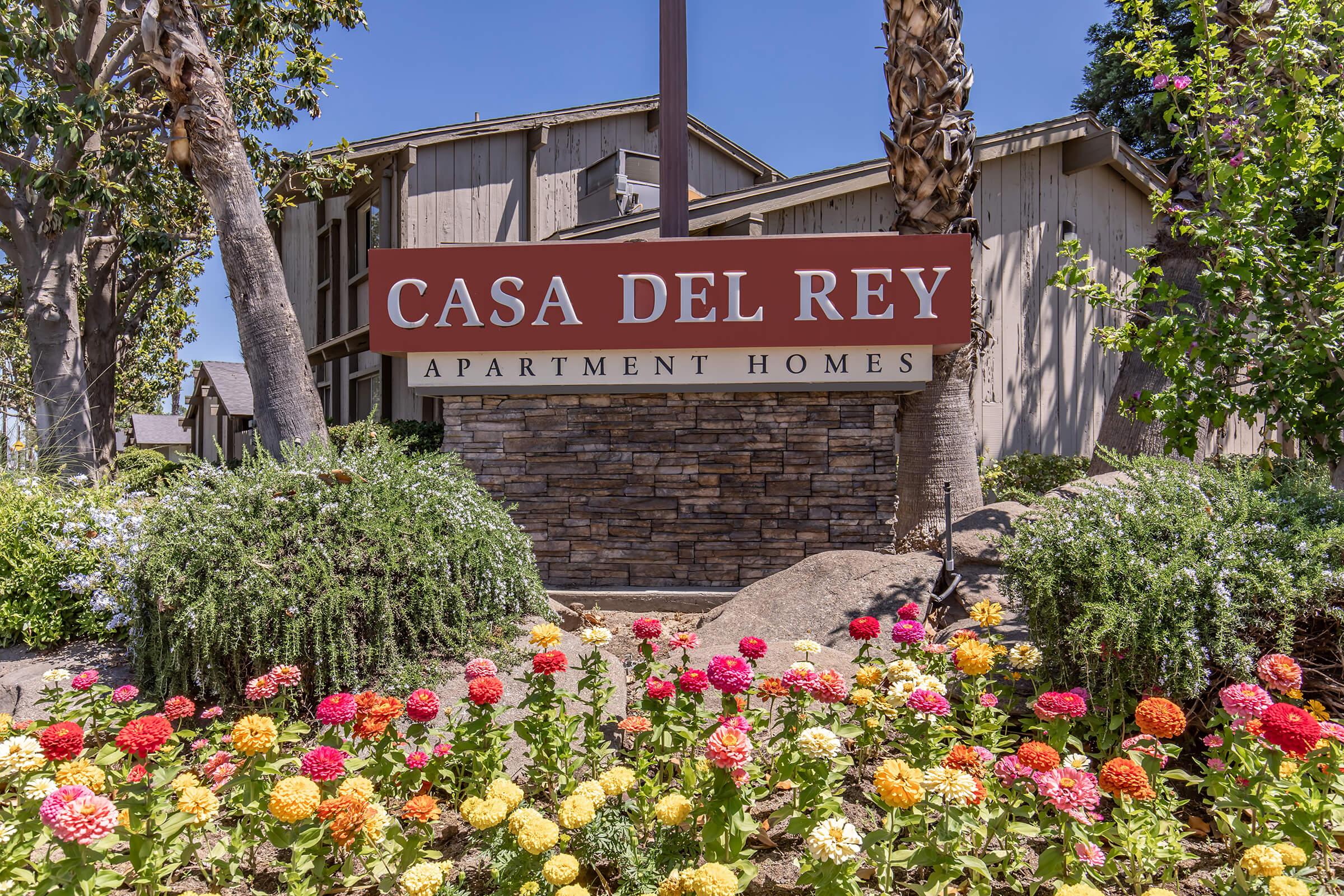Sign for Casa Del Rey Apartment Homes, featuring the name prominently displayed in red letters against a stone background. Surrounding the sign are vibrant flower beds with various colorful blooms, along with palm trees and a well-maintained landscape, contributing to a welcoming atmosphere.