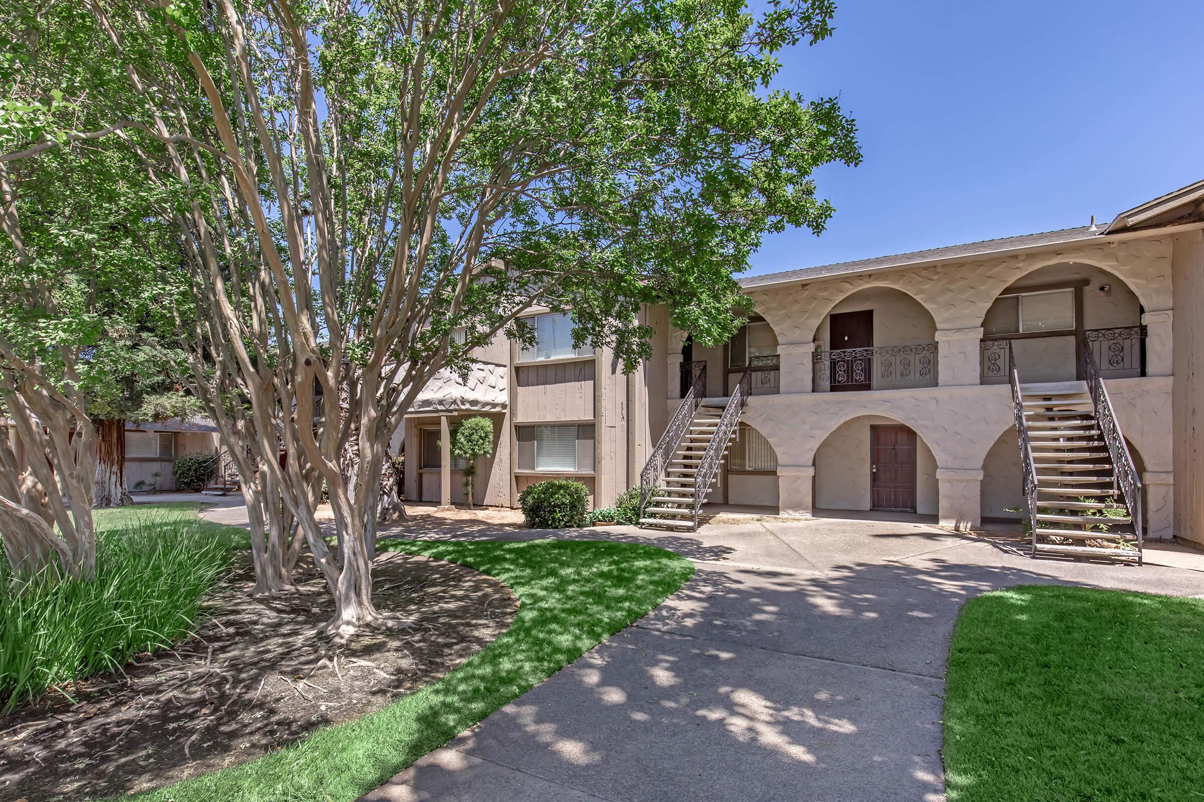 A view of a two-story apartment building with a courtyard. The building features archways and balconies. Lush green trees and shrubs surround the walkway, which is made of concrete. Bright blue sky is visible above, creating a pleasant outdoor atmosphere.