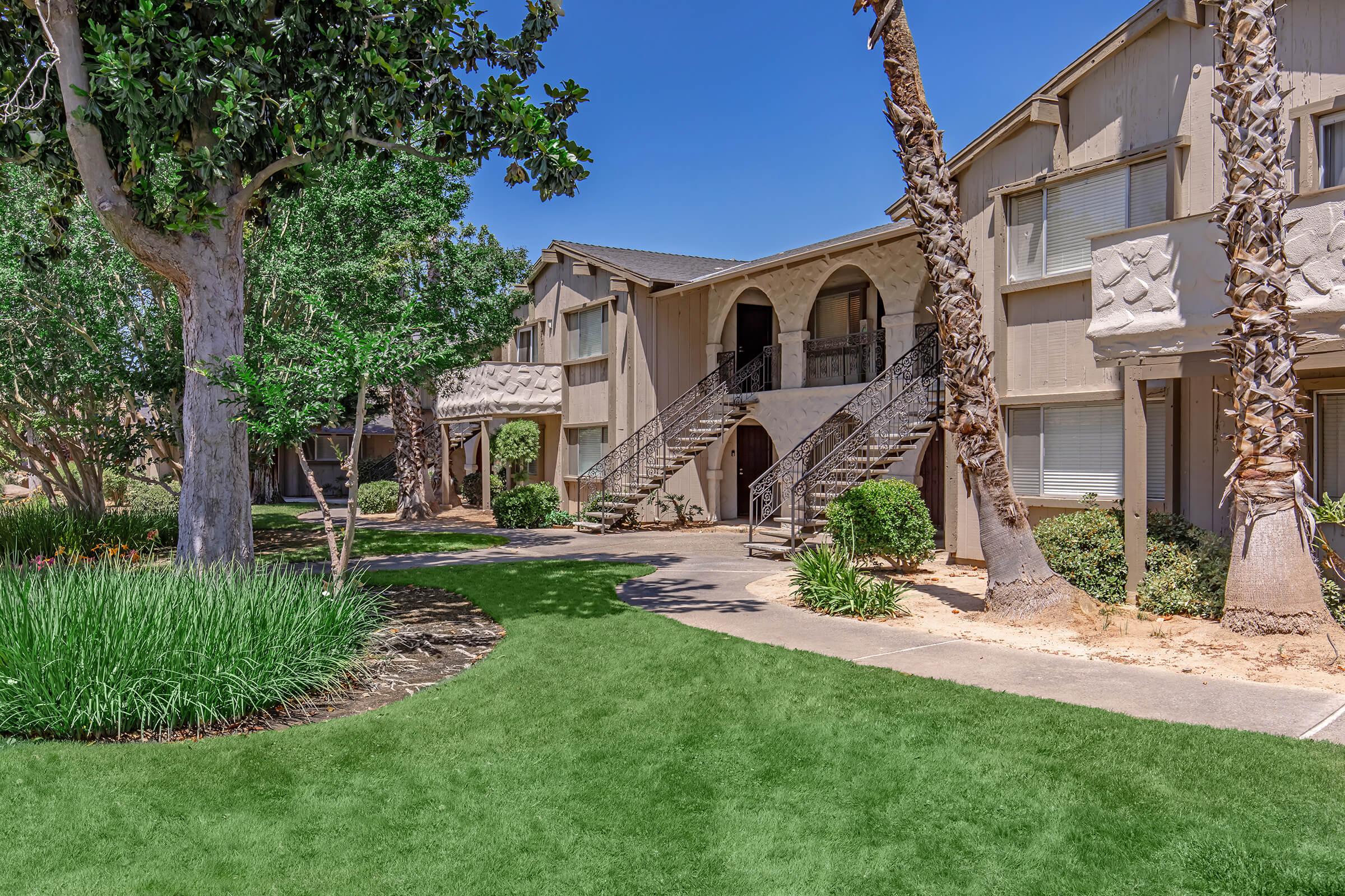 A well-maintained outdoor area featuring residential buildings with balconies, surrounded by lush green grass and palm trees. A pathway leads to the entrances, and the sky is clear and blue, creating a pleasant and inviting atmosphere.