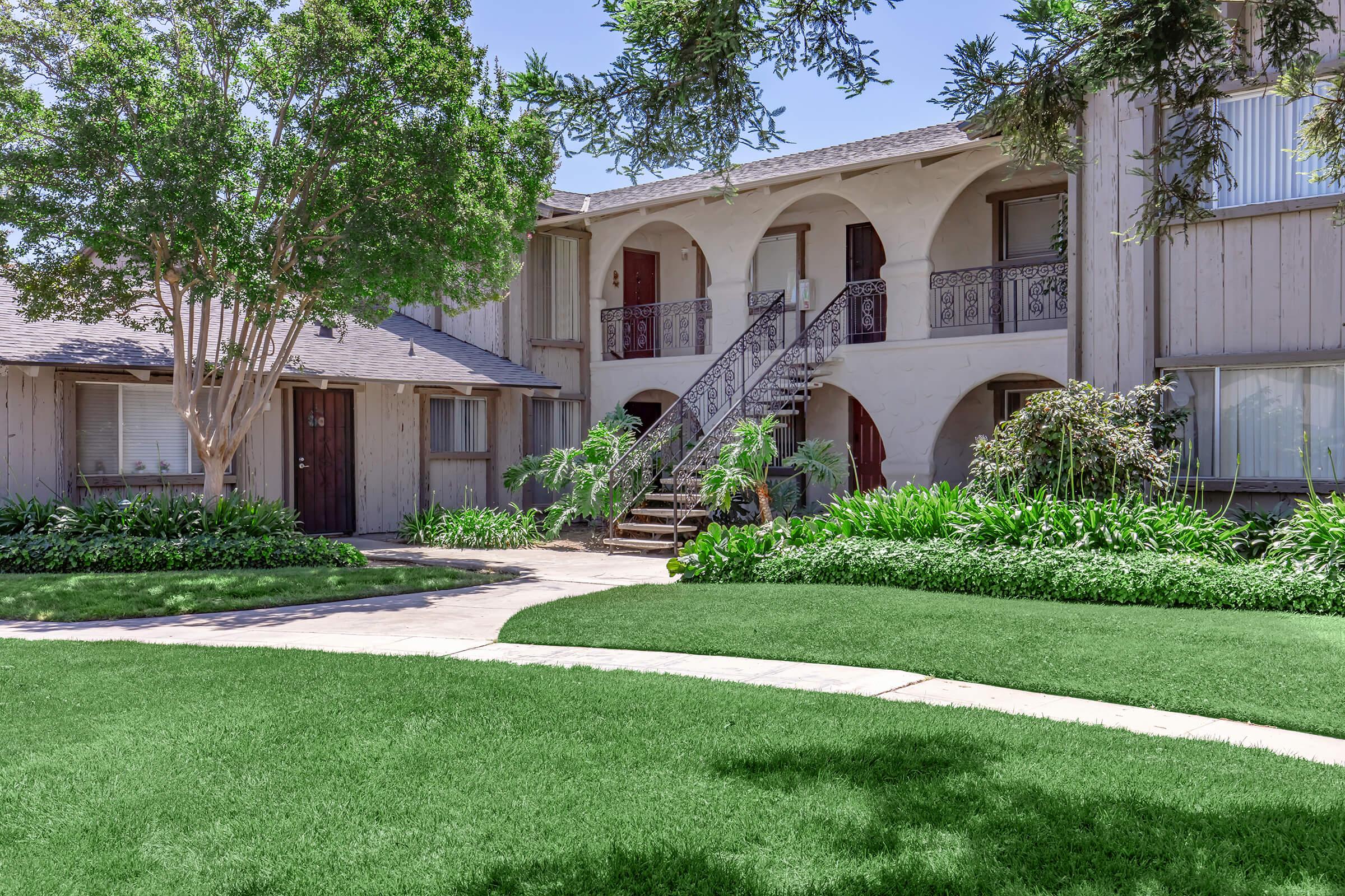 A low-rise apartment building featuring arched entrances, wooden paneling, and a central staircase surrounded by lush greenery and manicured lawns. The scene is bright and sunny, with trees providing shade to the area. The layout includes well-defined paths and flower beds.