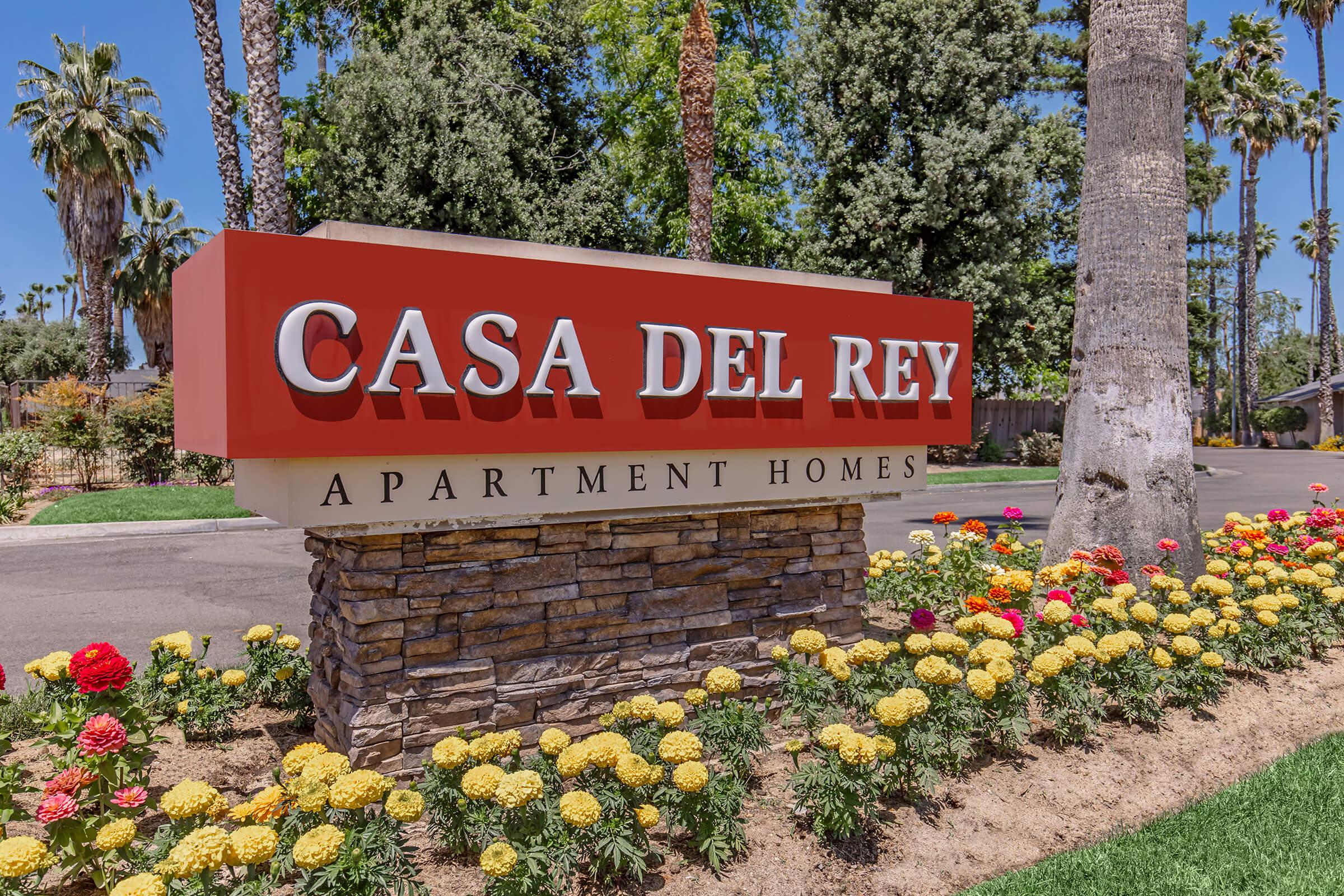 Sign for "Casa Del Rey Apartment Homes" prominently displayed, featuring red lettering against a stone base. Surrounded by colorful flower beds and palm trees, the setting is sunny and inviting, indicating a residential area.