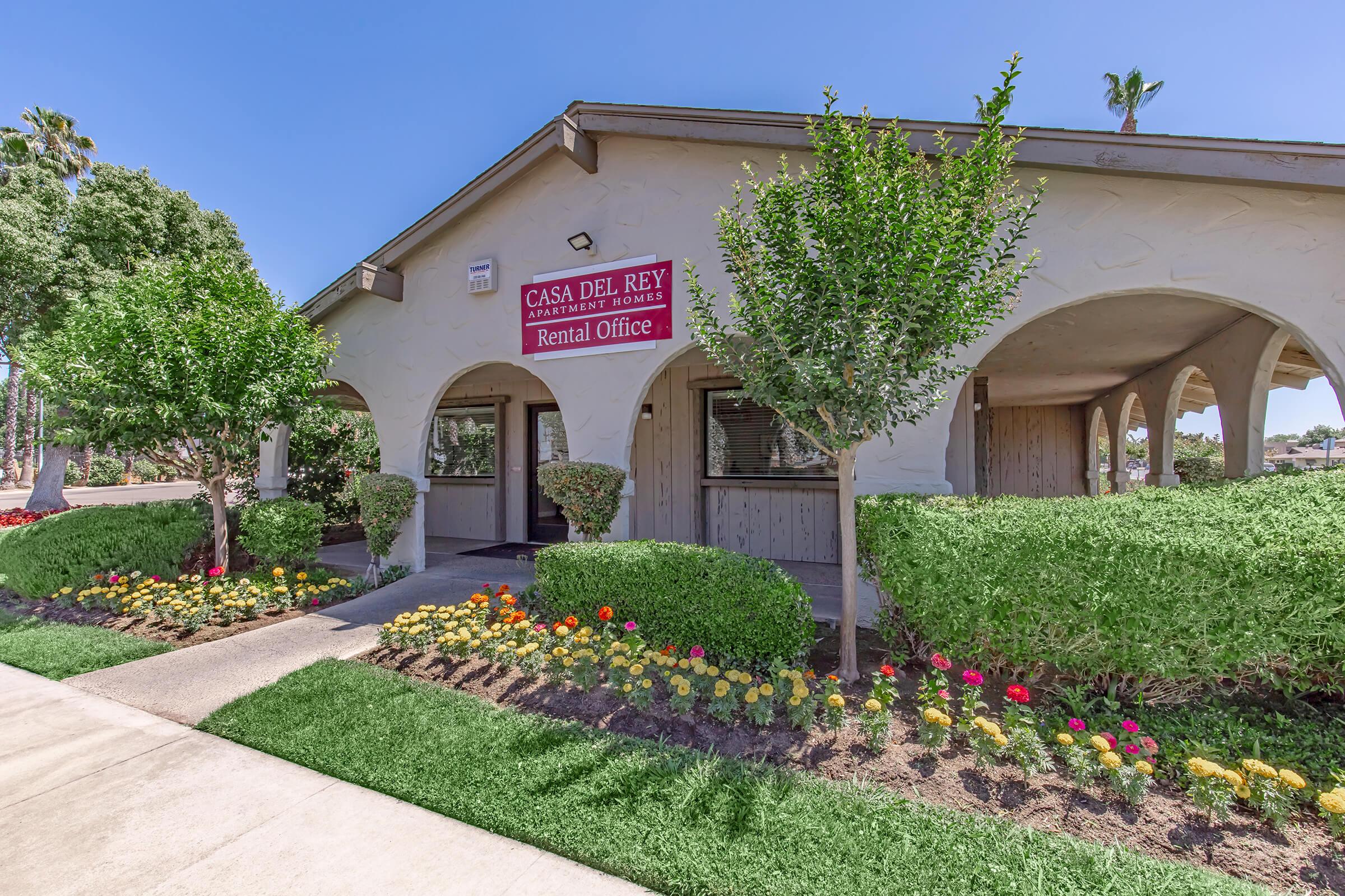 A rental office building named "Casa Del Rey" with a red sign. The structure features arched doorways and is surrounded by well-maintained landscaping with blooming flowers and green shrubs. The walkway leads up to the entrance, and palm trees are visible in the background against a clear blue sky.