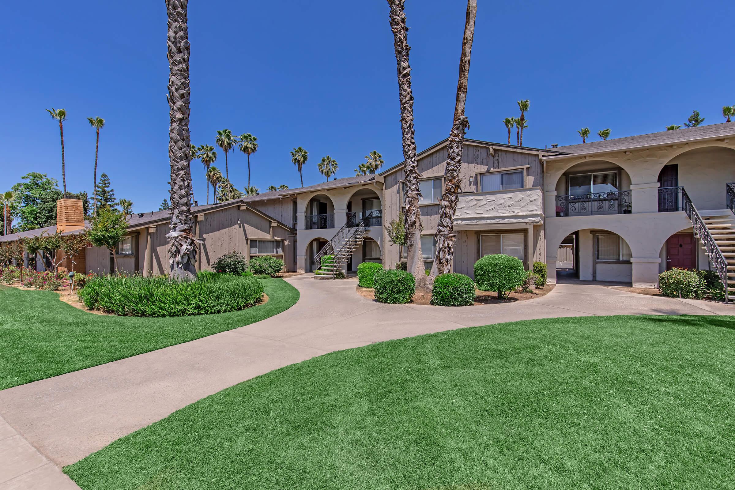 A landscaped courtyard featuring two-story apartment buildings surrounded by palm trees and manicured greenery. A paved walkway leads through the lawn, connecting different sections of the complex, under a clear blue sky. The architecture includes arched entries and balconies.