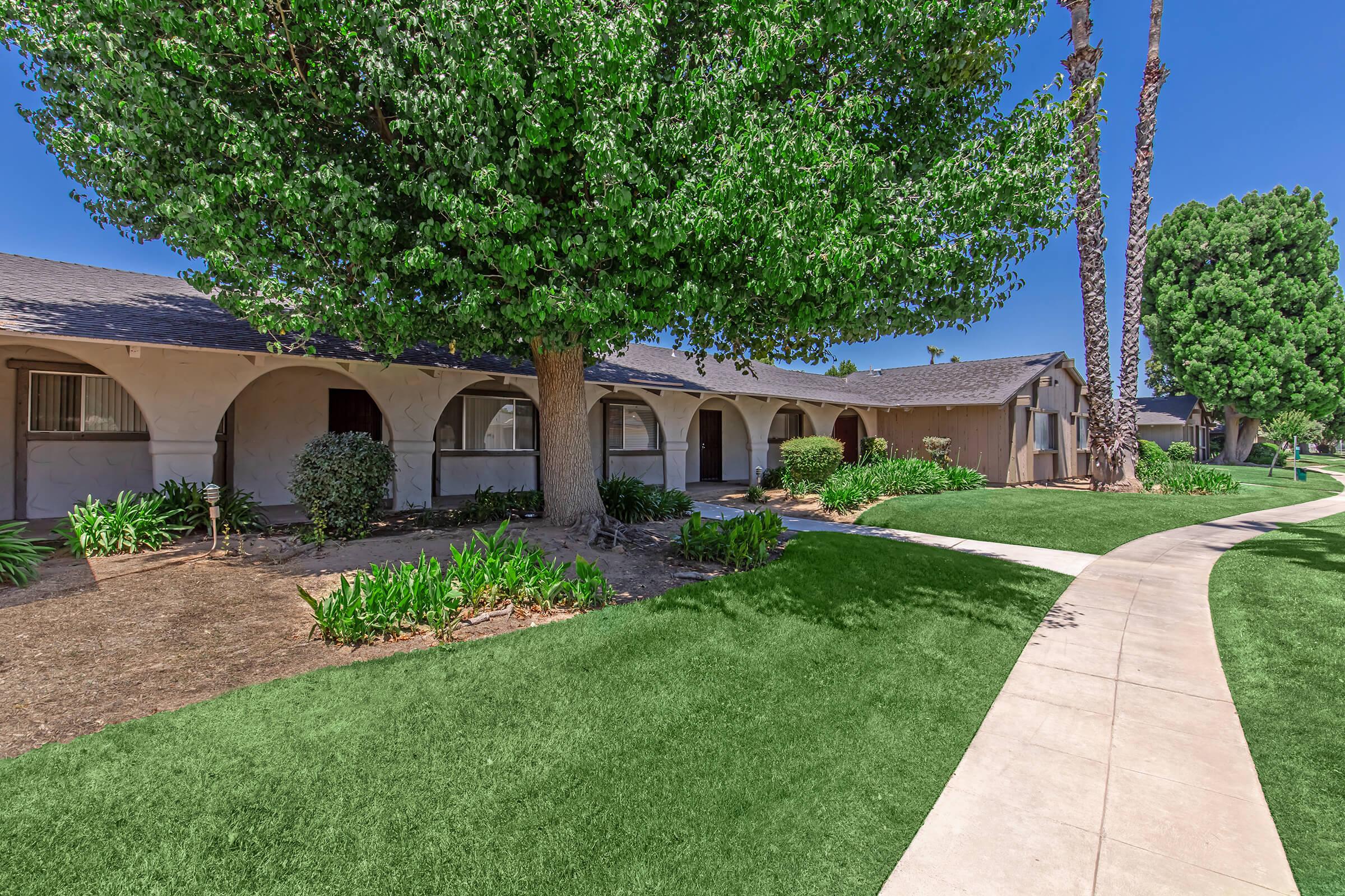 A landscaped walkway leads through a green yard with vibrant plants and trees in front of a series of residential buildings featuring arched entrances. The sky is clear blue, enhancing the inviting atmosphere of the area.