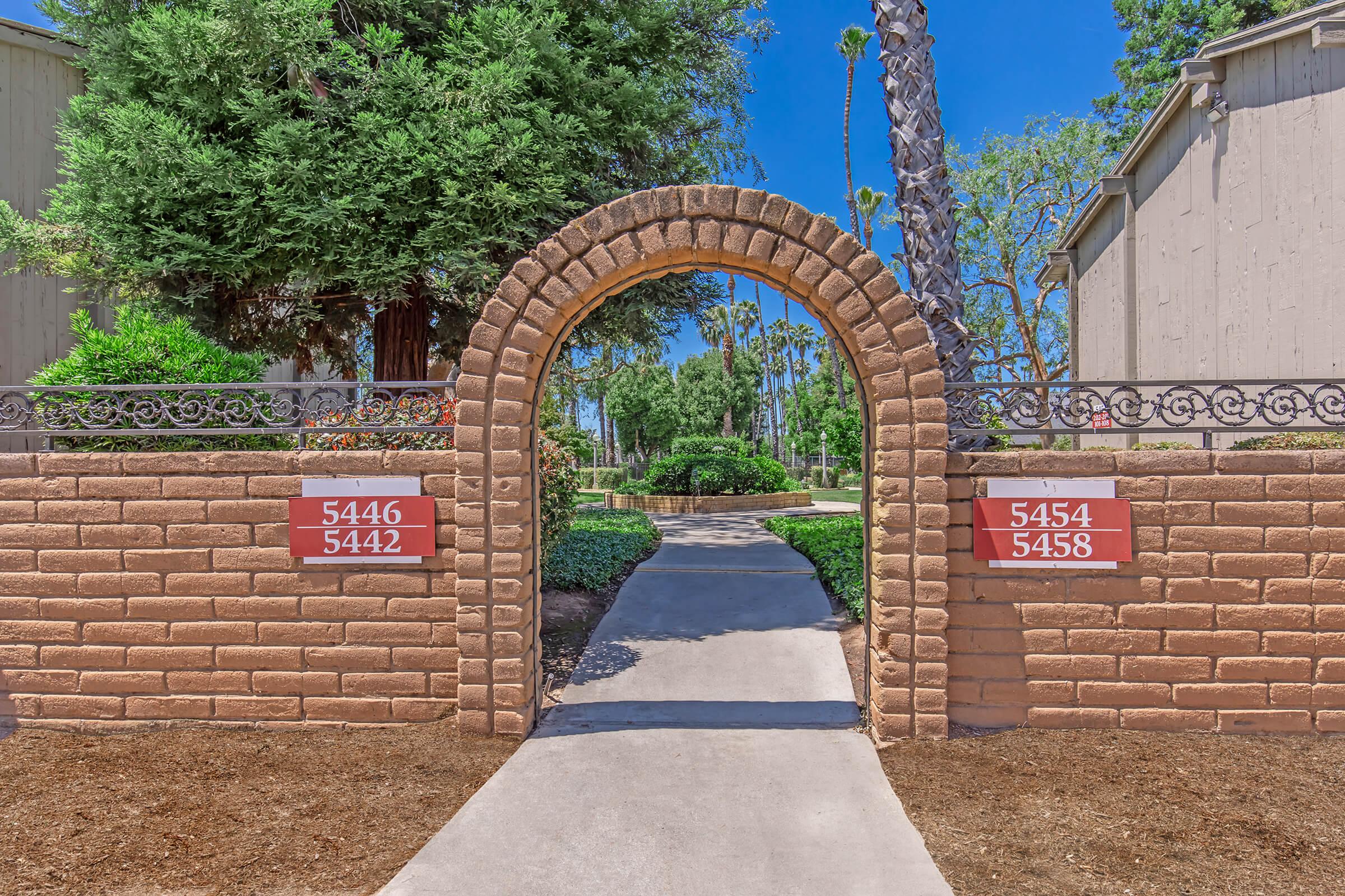 An arched stone entrance with red number plaques reading "5442" on the left and "5458" on the right, leading to a well-maintained walkway surrounded by greenery, including trees and shrubs, under a clear blue sky.