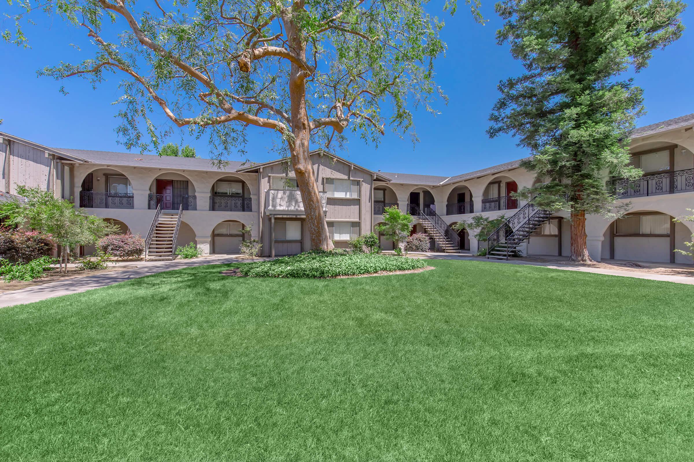 A large, green courtyard surrounded by two-story apartment buildings. The courtyard features manicured grass, a mature tree, and decorative shrubs. There are staircases leading to the upper levels of the buildings, with clear blue skies in the background.