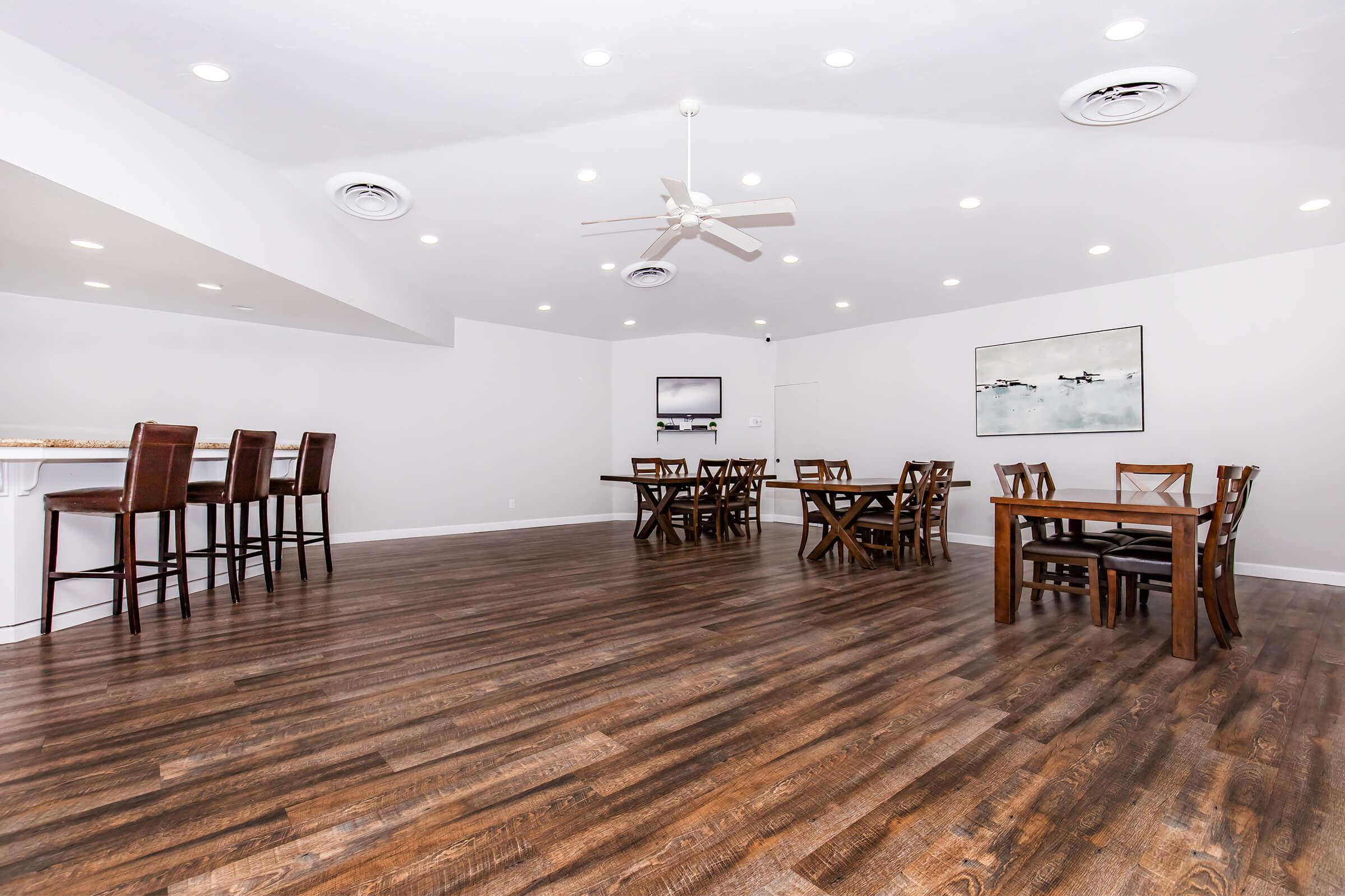 A spacious, modern room featuring a polished wooden floor. There are several wooden tables with chairs arranged throughout the space. A small bar area with high stools is on one side. The walls are painted white, and there is a ceiling fan and recessed lighting, creating a bright and inviting atmosphere.