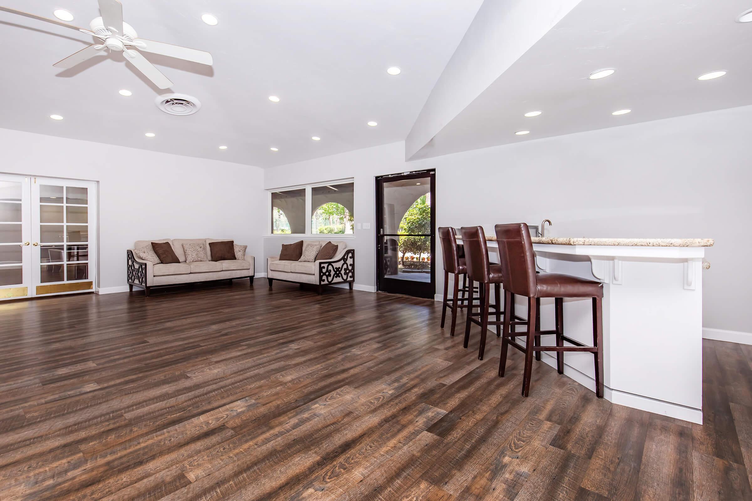 Bright and spacious living area featuring hardwood floors, two sofas, and a bar with high stools. Large windows allow natural light to fill the space, and double doors lead to an outdoor area. The ceiling is accentuated by recessed lighting and a ceiling fan for added comfort.