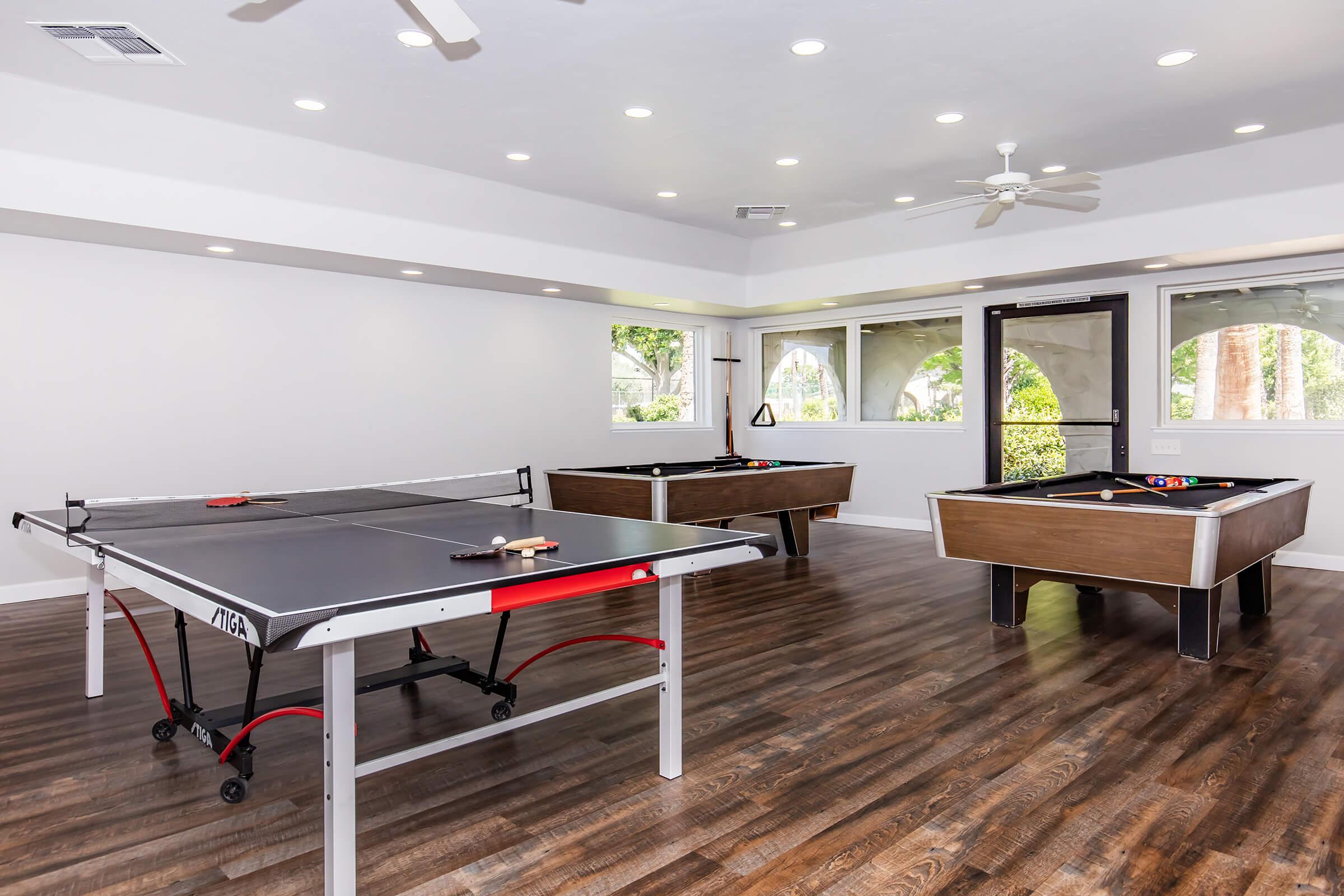 A recreational room featuring two tables: a black ping pong table and a wooden pool table. The space has hardwood floors and large windows providing natural light, with a ceiling fan for ventilation. The walls are painted in a light color, creating a spacious and inviting atmosphere for games.