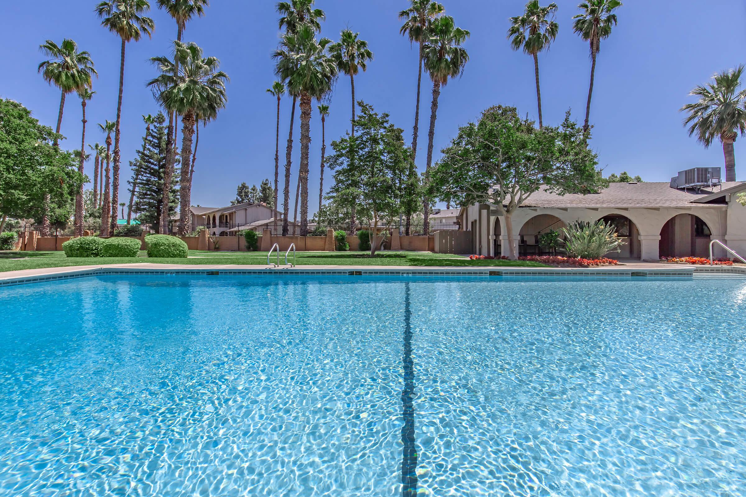 A serene swimming pool surrounded by tall palm trees and lush green landscaping. The clear blue water reflects the sunlight, creating a tranquil atmosphere. In the background, a well-maintained building with arches and vibrant flower beds adds to the inviting scenery. Perfect for relaxation and leisure.