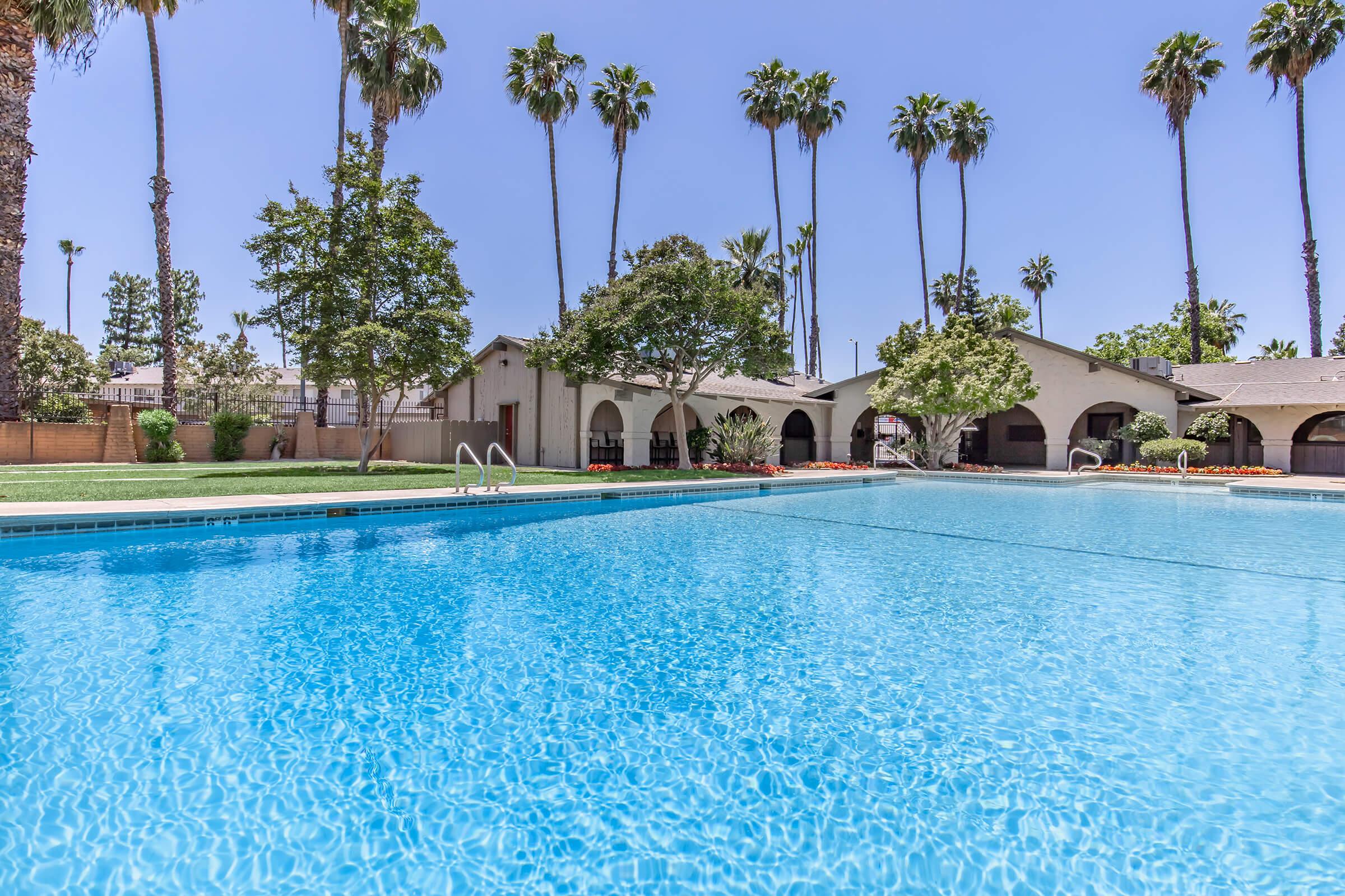 A serene swimming pool surrounded by palm trees and lush greenery, with a clear blue sky overhead. The pool features a well-maintained surface reflecting the sunlight, and there are buildings in the background with arched architecture. Perfect for relaxation and enjoying a sunny day.