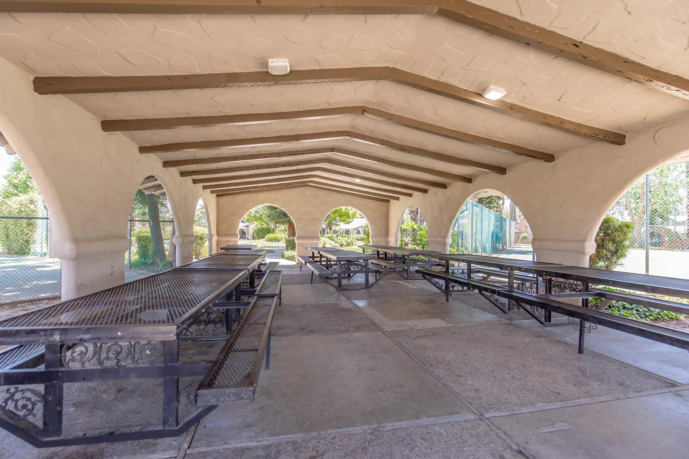 A spacious, covered outdoor area featuring several metal picnic tables arranged in rows. The structure has a high, arching ceiling with wooden beams and a stone-textured wall. Natural light filters through the arches, surrounded by greenery and trees, creating a serene setting.