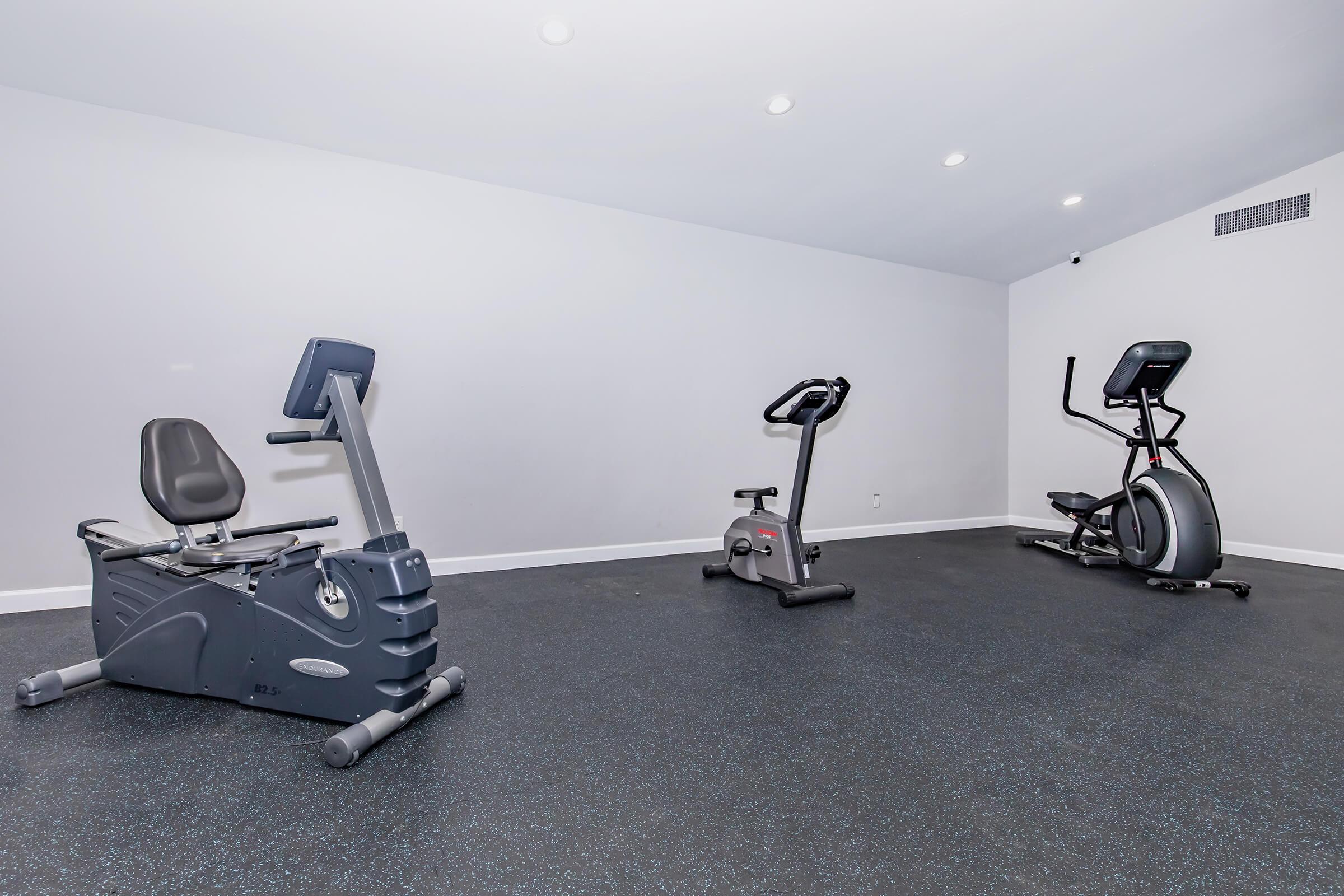 A modern home gym featuring a stationary bike, an elliptical machine, and a recumbent bike. The walls are white, and the floor has a dark rubber surface, with recessed lighting above. The room is bright and minimalistic, designed for workouts.