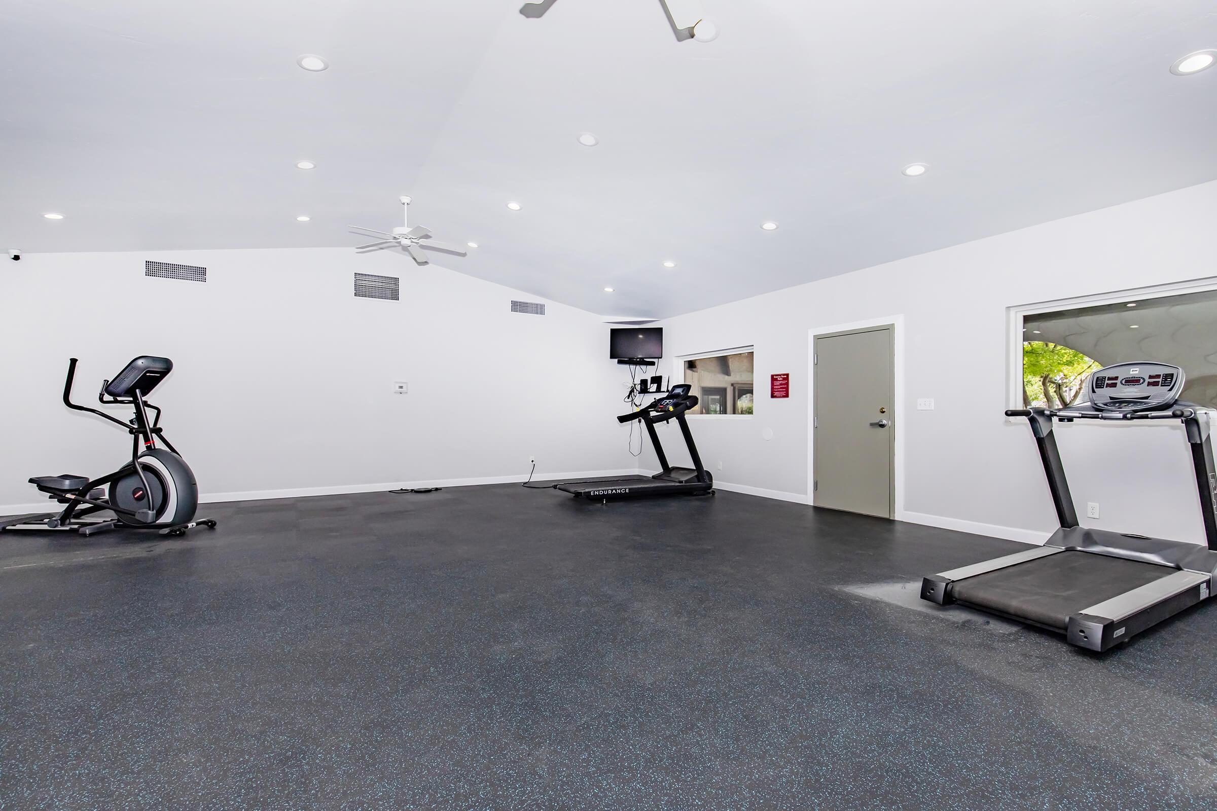 A spacious, well-lit gym featuring exercise equipment including an elliptical machine, a treadmill, and a stationary bike. The room has a ceiling fan, recessed lighting, and large windows, enhancing the open atmosphere. A wall-mounted TV is visible, and the flooring is dark rubber.