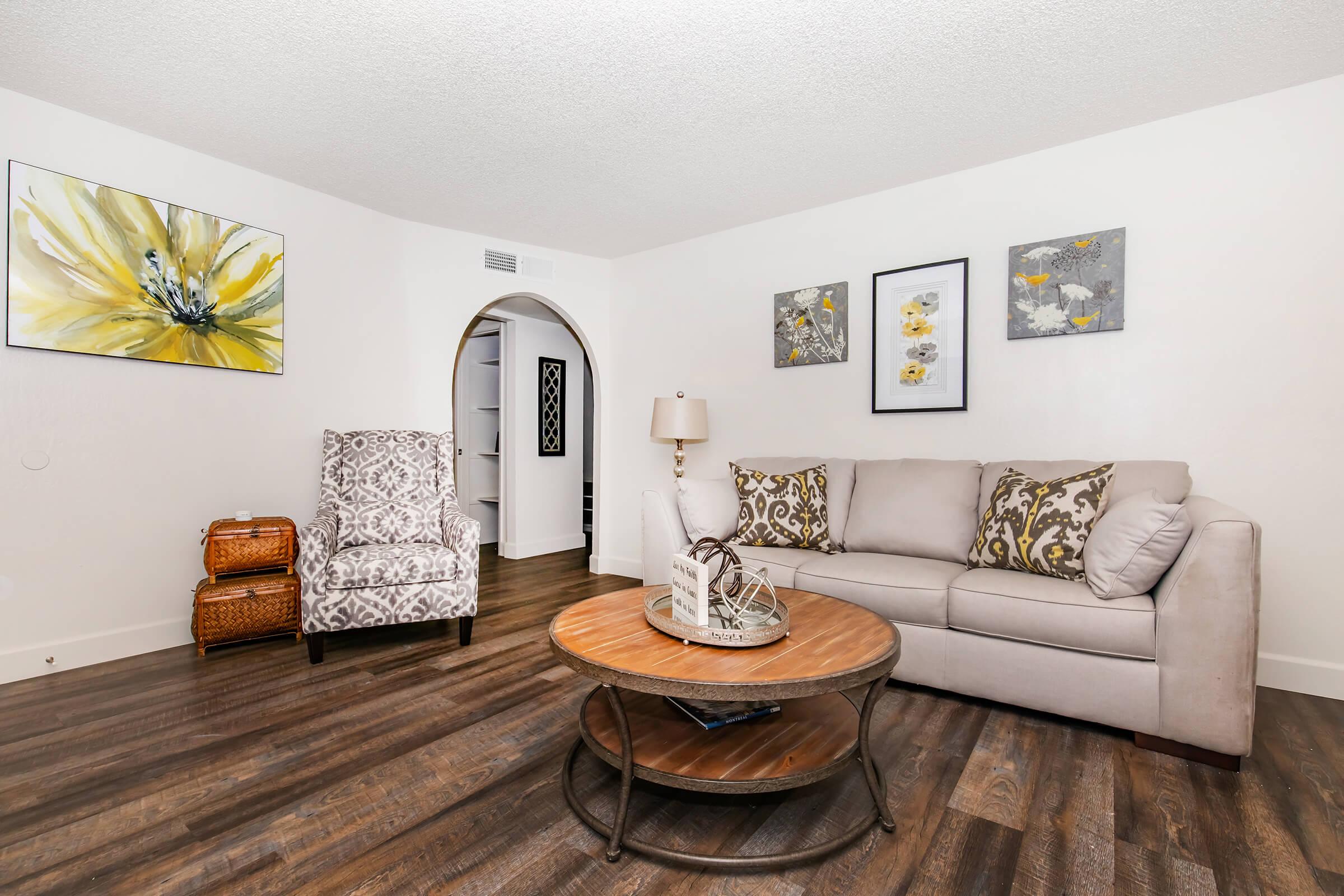 A cozy living room featuring a light-colored sofa with decorative pillows, a round wooden coffee table, and a patterned armchair. The walls are adorned with floral art, and there are two small baskets in the corner. The flooring is dark wood, creating a warm ambiance. A doorway leads to another space in the background.