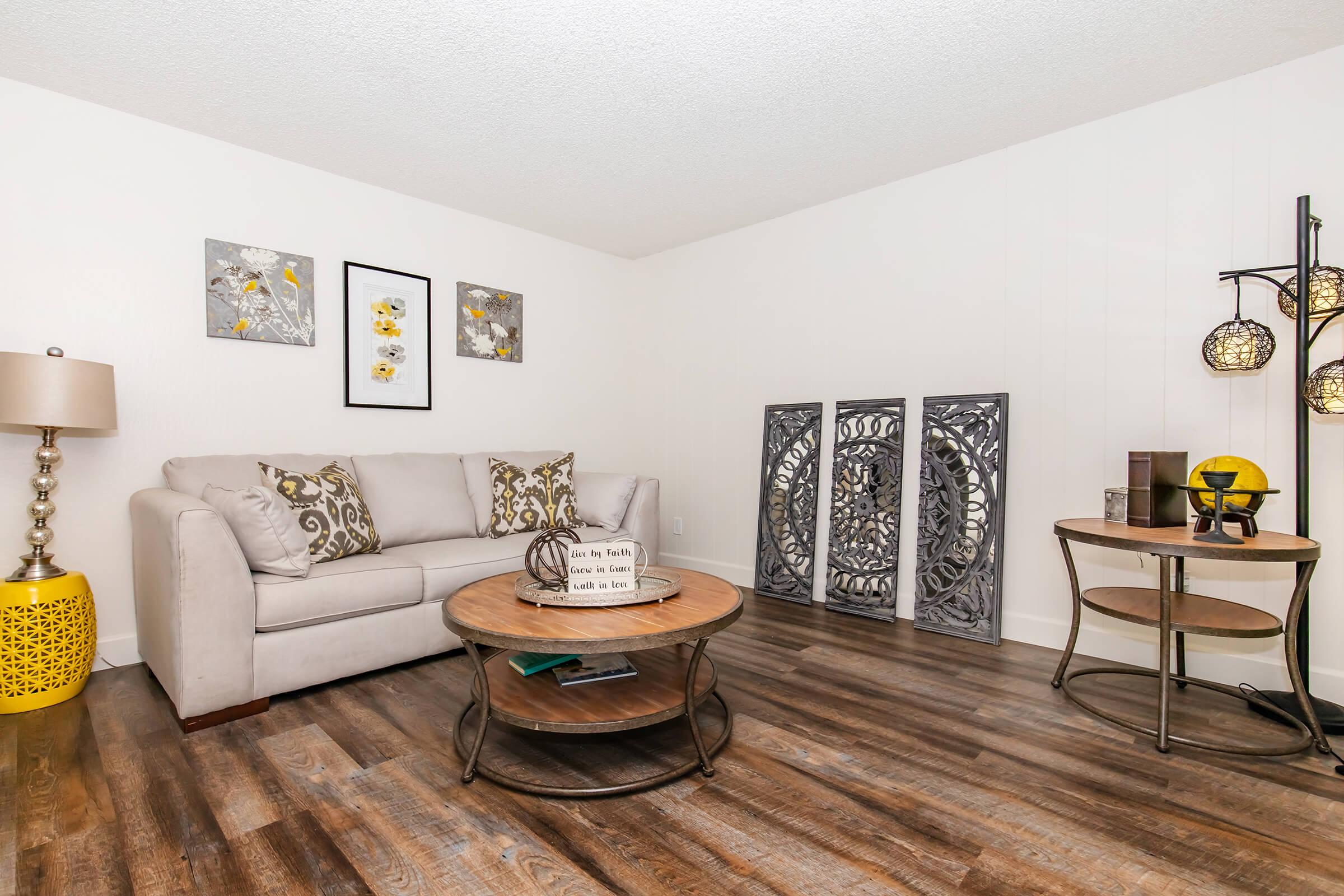 A cozy living room featuring a light gray sofa adorned with decorative pillows, a round wooden coffee table, and a decorative floor lamp. On the walls, there are framed art pieces and an intricate metal wall decoration. A small circular table is set up with a decorative item, enhancing the inviting atmosphere.