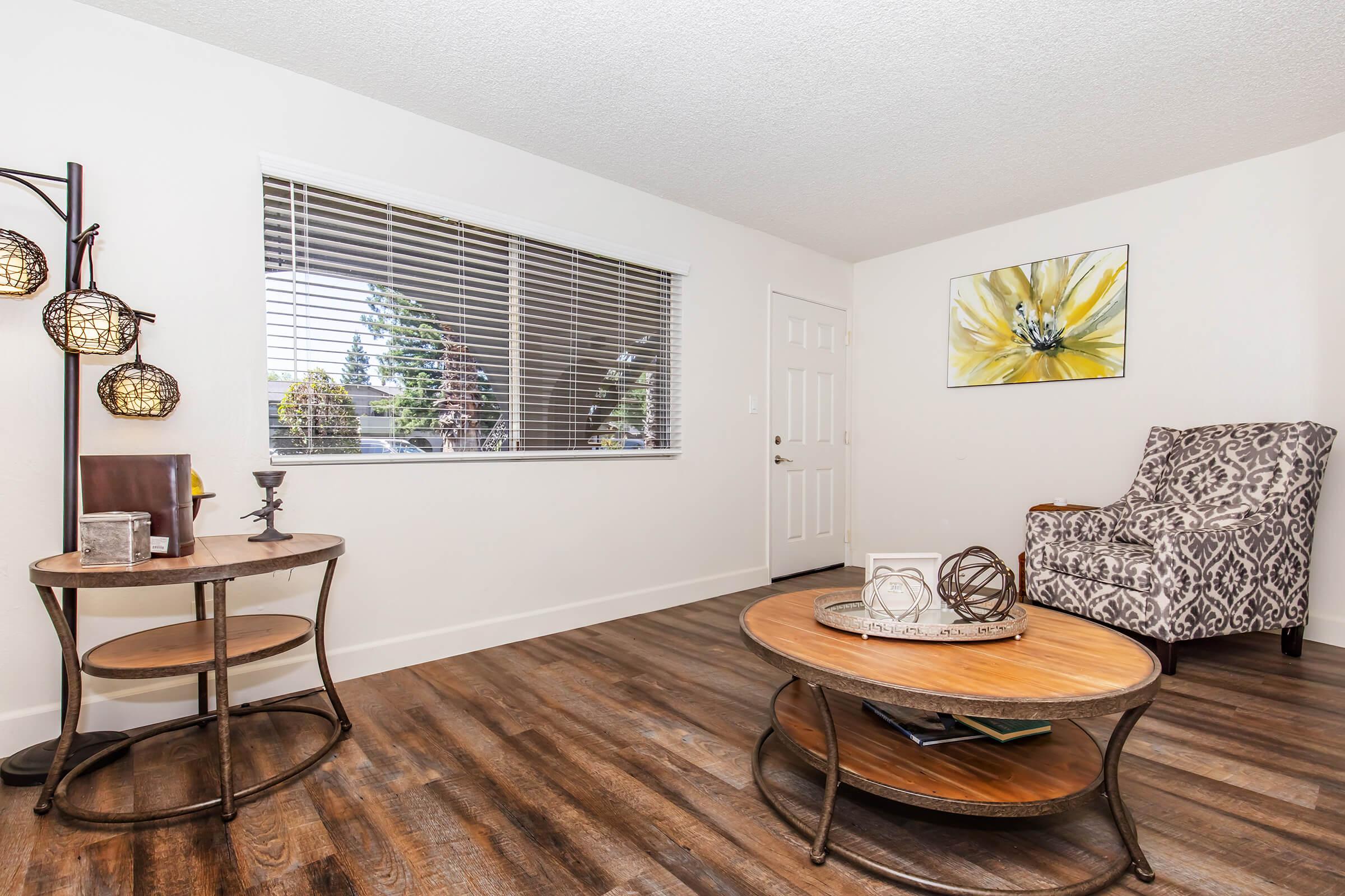 A cozy living room featuring a patterned armchair, a round wooden coffee table, and a side table with decorative items. The walls are painted white, and a large window with blinds allows natural light to enter. A floral artwork adds a touch of color to the space. The flooring is wooden, enhancing the warm ambiance.