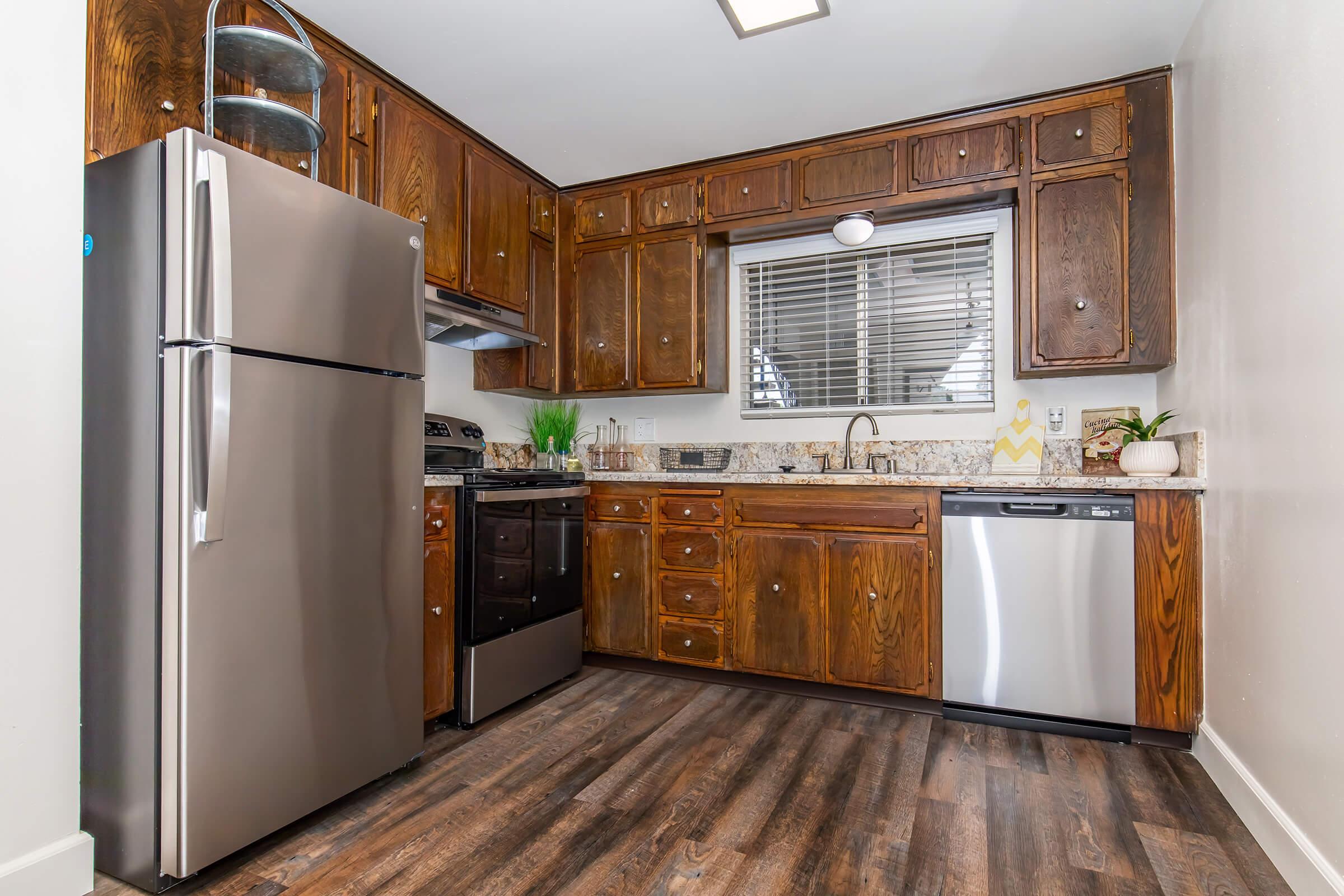 A modern kitchen featuring dark wooden cabinets and stainless steel appliances. The space includes a refrigerator, oven, microwave, dishwasher, and a sink with a window above it. The countertop is made of granite and there are decorative plants on the counter. The flooring is dark wood laminate.