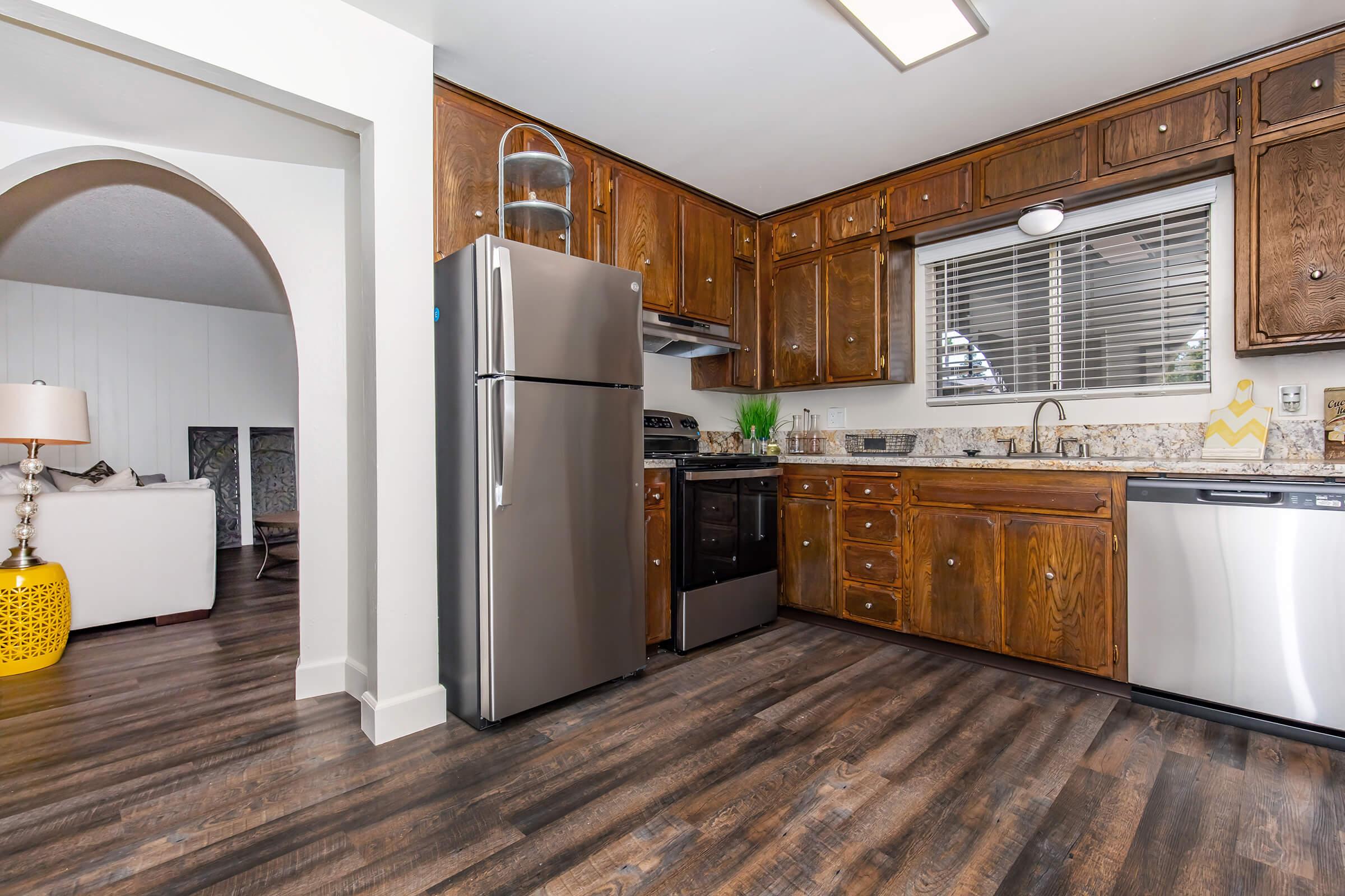 A modern kitchen featuring dark wood cabinets, stainless steel appliances including a refrigerator and oven, a granite countertop, and a window with blinds. The flooring is dark wood, and a glimpse of a living area with a white couch and decorative yellow accent table can be seen through an archway.