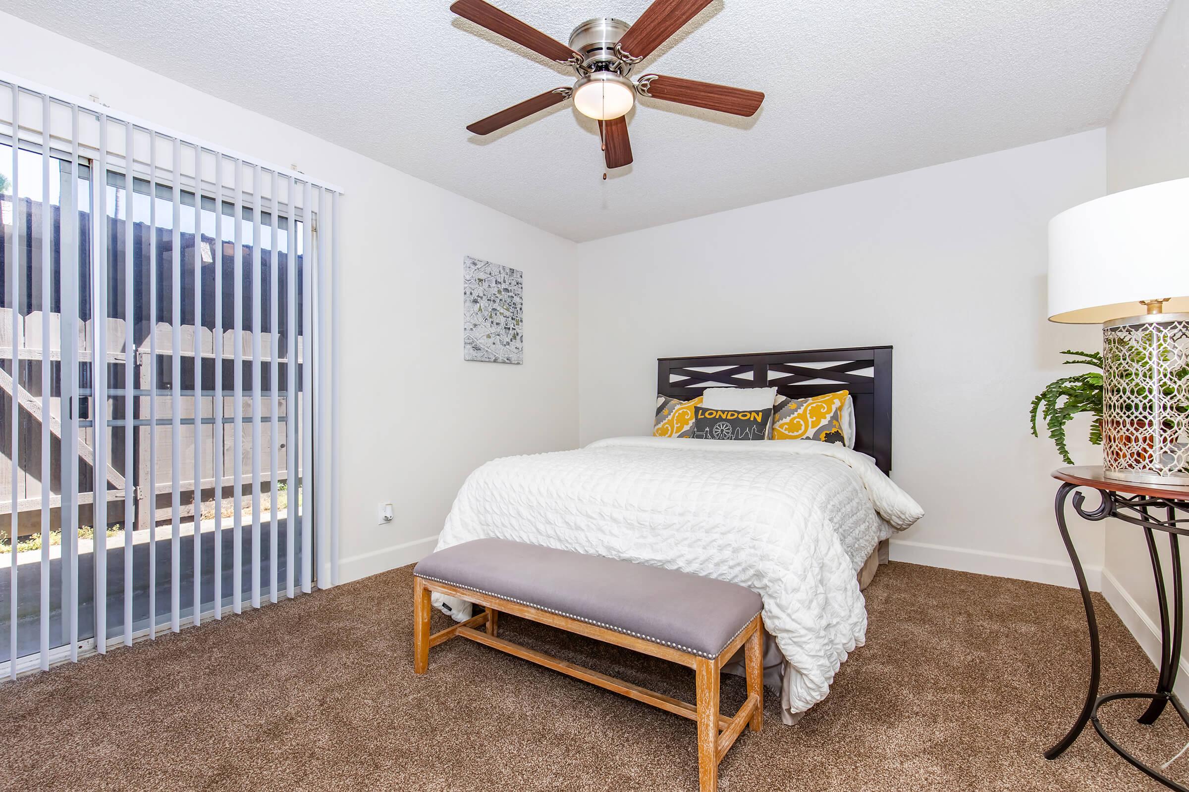 A cozy bedroom featuring a queen-sized bed with decorative pillows, a bench at the foot of the bed, a ceiling fan, and a window with vertical blinds. The room has carpeted flooring and a lamp on a side table, creating a warm and inviting atmosphere.