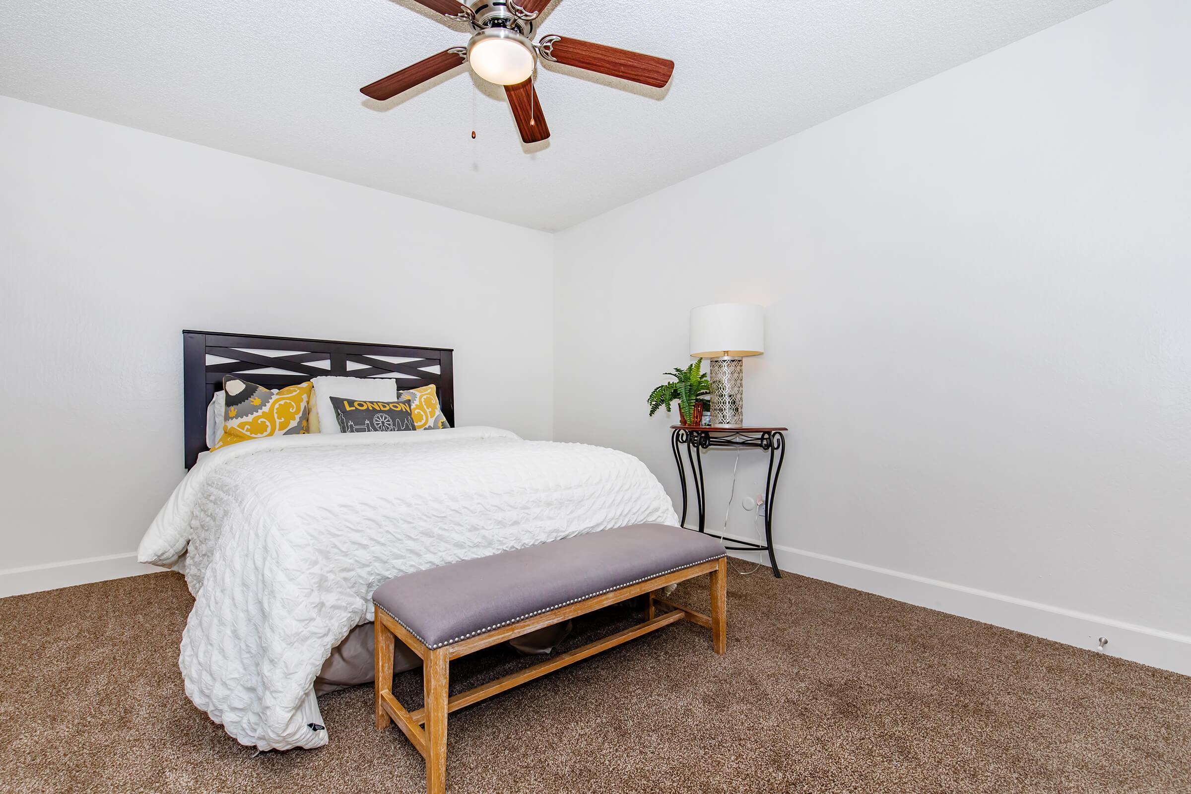 A cozy bedroom featuring a queen-sized bed with a white quilt and yellow accent pillows, a decorative bench at the foot of the bed, a stylish lamp on a small side table, and a ceiling fan. The room has light-colored walls and soft brown carpet, creating a warm and inviting atmosphere.