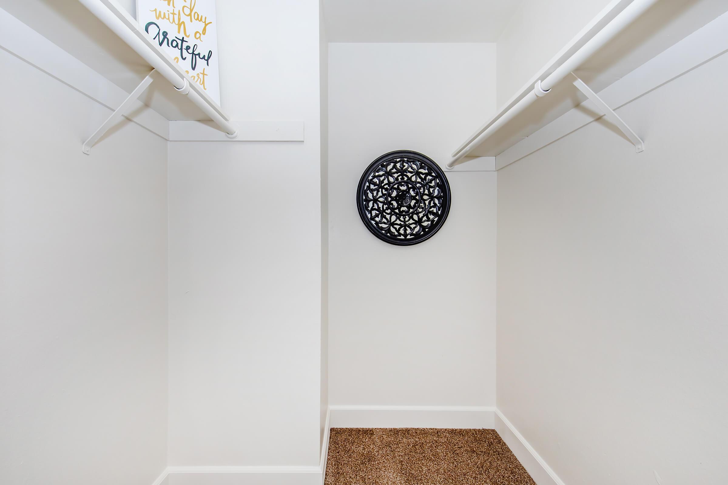 A spacious, empty closet featuring white walls and a tan carpet. Two hanging rods are installed along the sides, and a decorative black circular wall piece is mounted on the back wall. A framed sign with golden text is visible at the top left, adding a touch of decor to the otherwise minimalistic space.