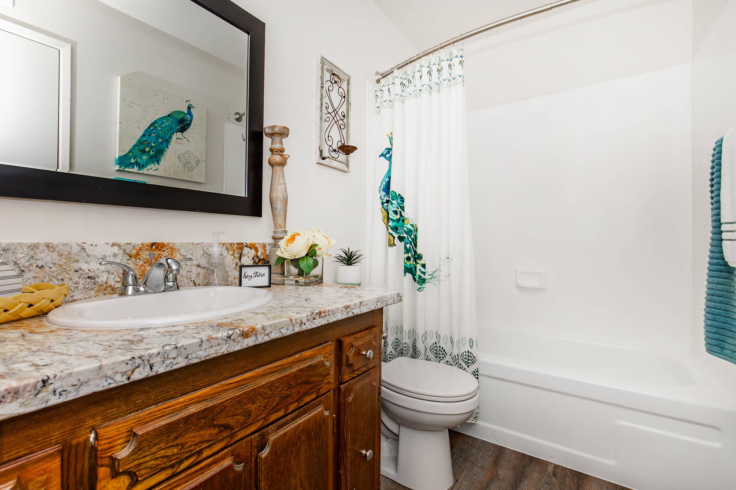 A modern bathroom featuring a granite countertop with a sink, wooden cabinetry, and a toilet. The bathtub is surrounded by a vibrant shower curtain with peacock designs. A decorative mirror, a candle holder, and potted plants add elegance to the space. Soft blue towels hang on the wall, enhancing the decor.