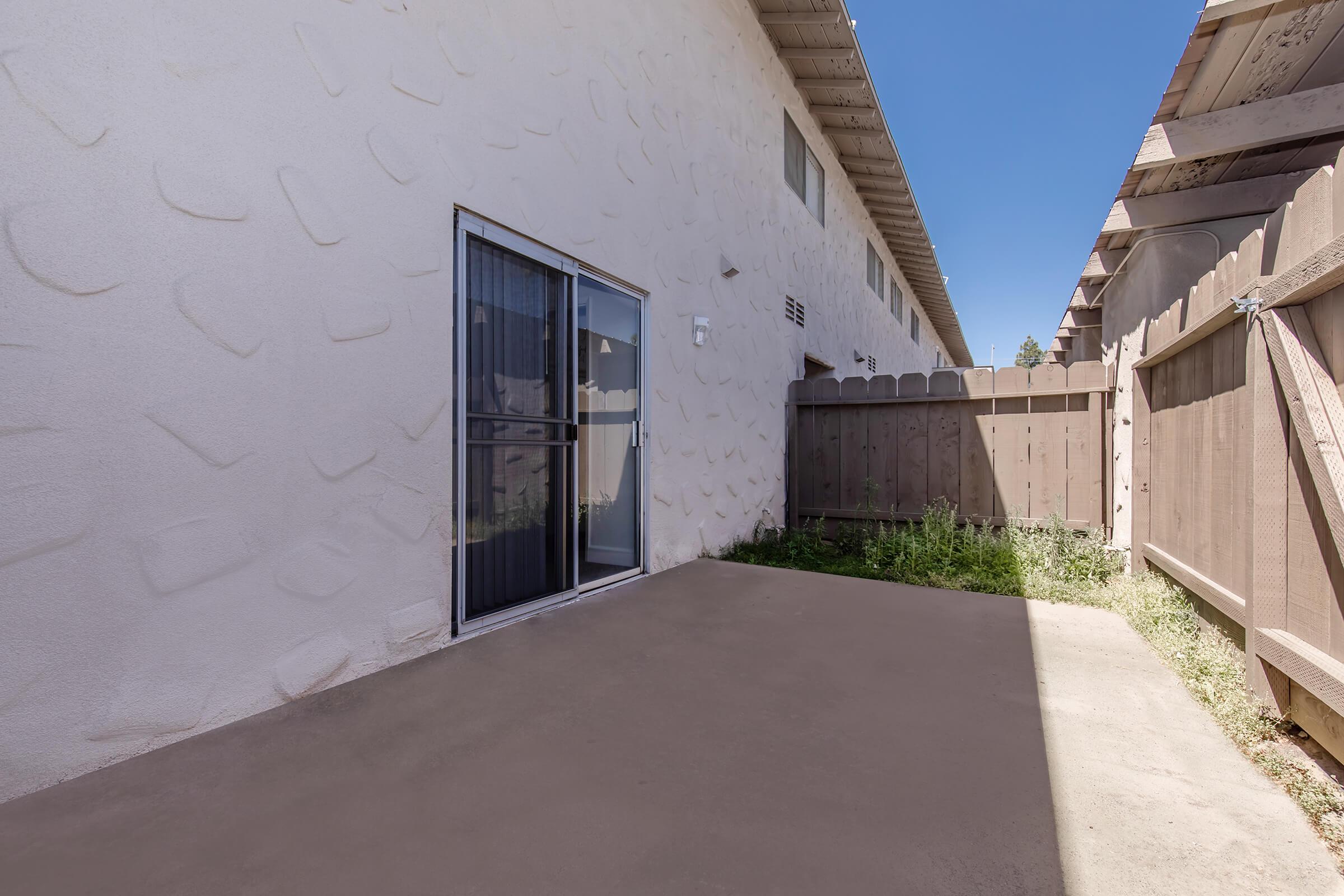 A small outdoor patio area next to a building, featuring a glass sliding door and a fenced perimeter. The ground is mostly concrete with some grass and weeds growing in a corner, and the wall of the adjacent building is textured and light-colored.