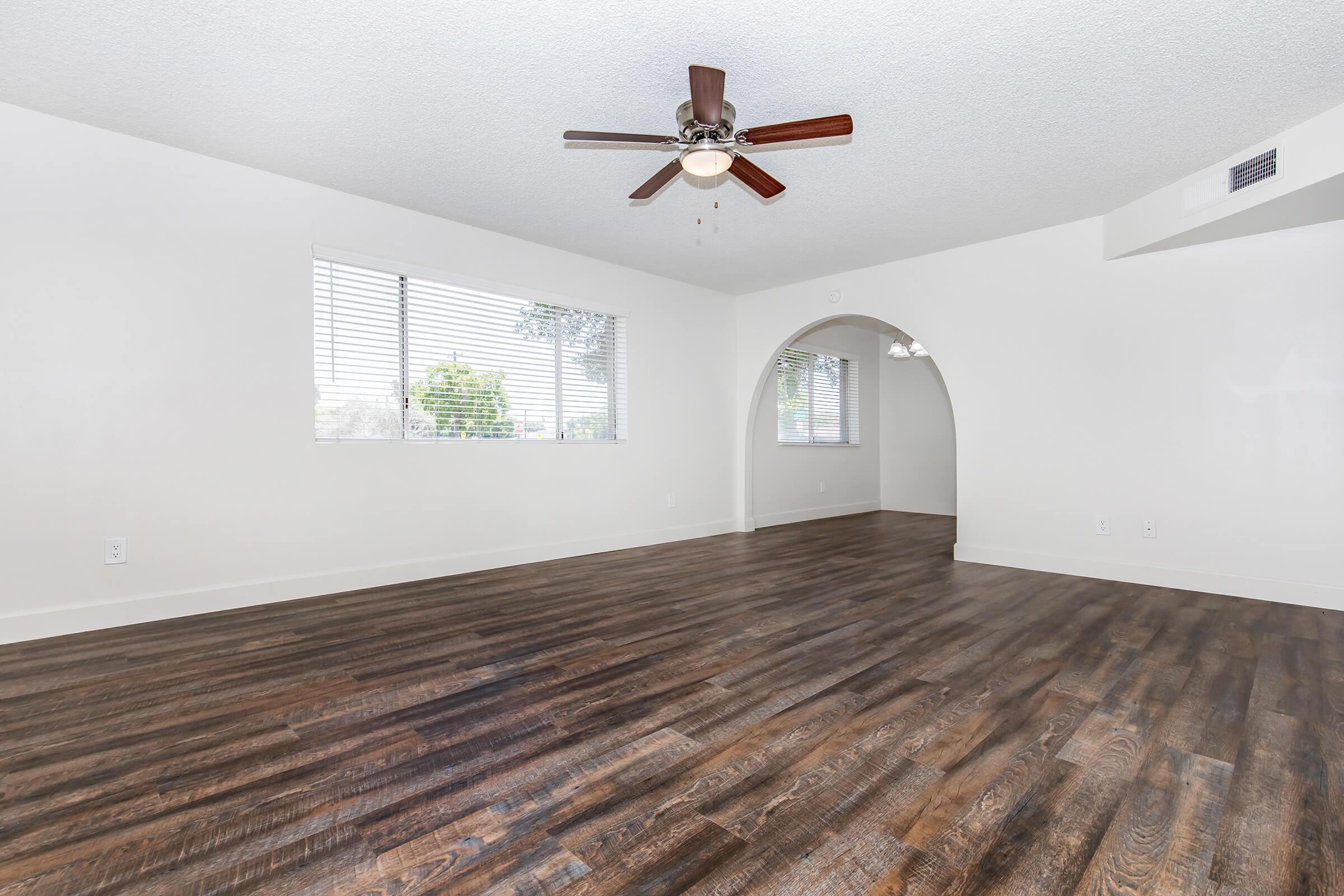 Spacious living room featuring laminate wood flooring, a ceiling fan with light fixture, and large windows providing natural light. An archway leads to another room, enhancing the open layout. The walls are painted a light color, creating a bright and inviting atmosphere.