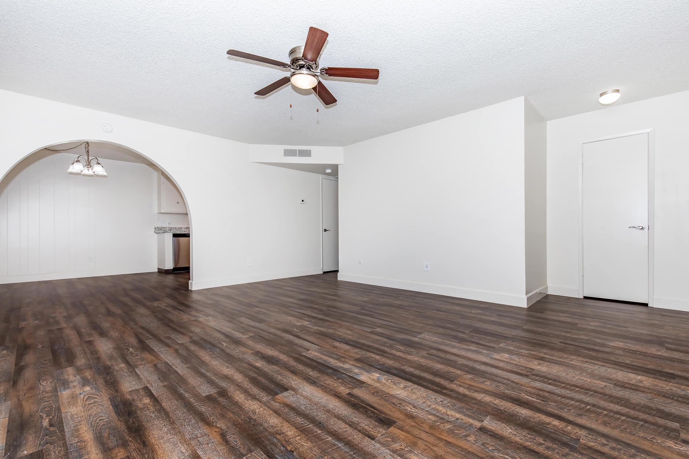 Spacious living area featuring a ceiling fan, light hardwood flooring, and a neutral color palette. An archway leads to a kitchen area in the background, with a door on the right leading to another room. The open layout offers plenty of natural light and space for furniture arrangements.