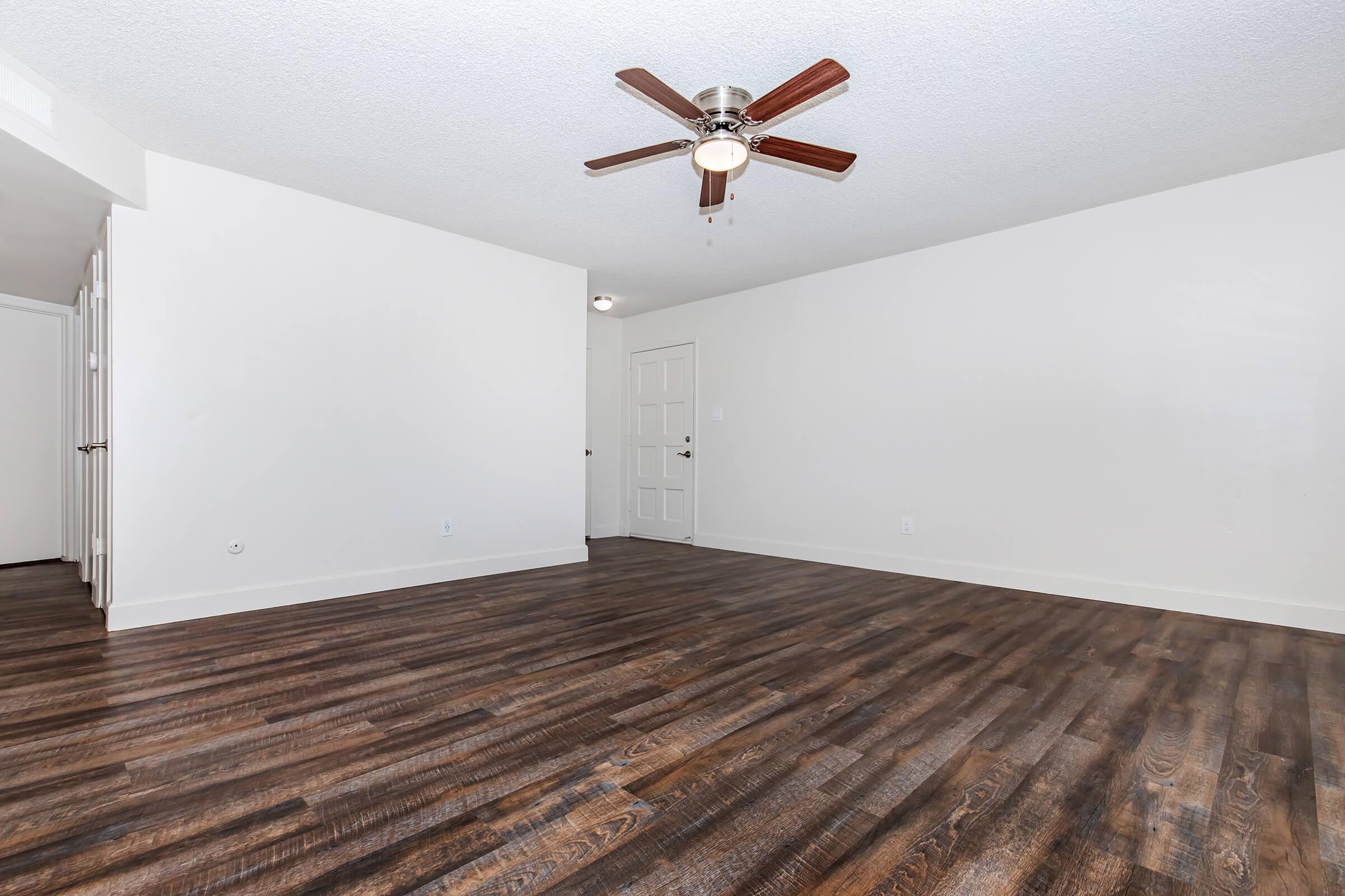 A spacious, unfurnished living room with dark wooden flooring, a ceiling fan with light, and white walls. There are doorways leading to other rooms, suggesting a bright and open layout. The room is well-lit and has a clean, modern appearance.