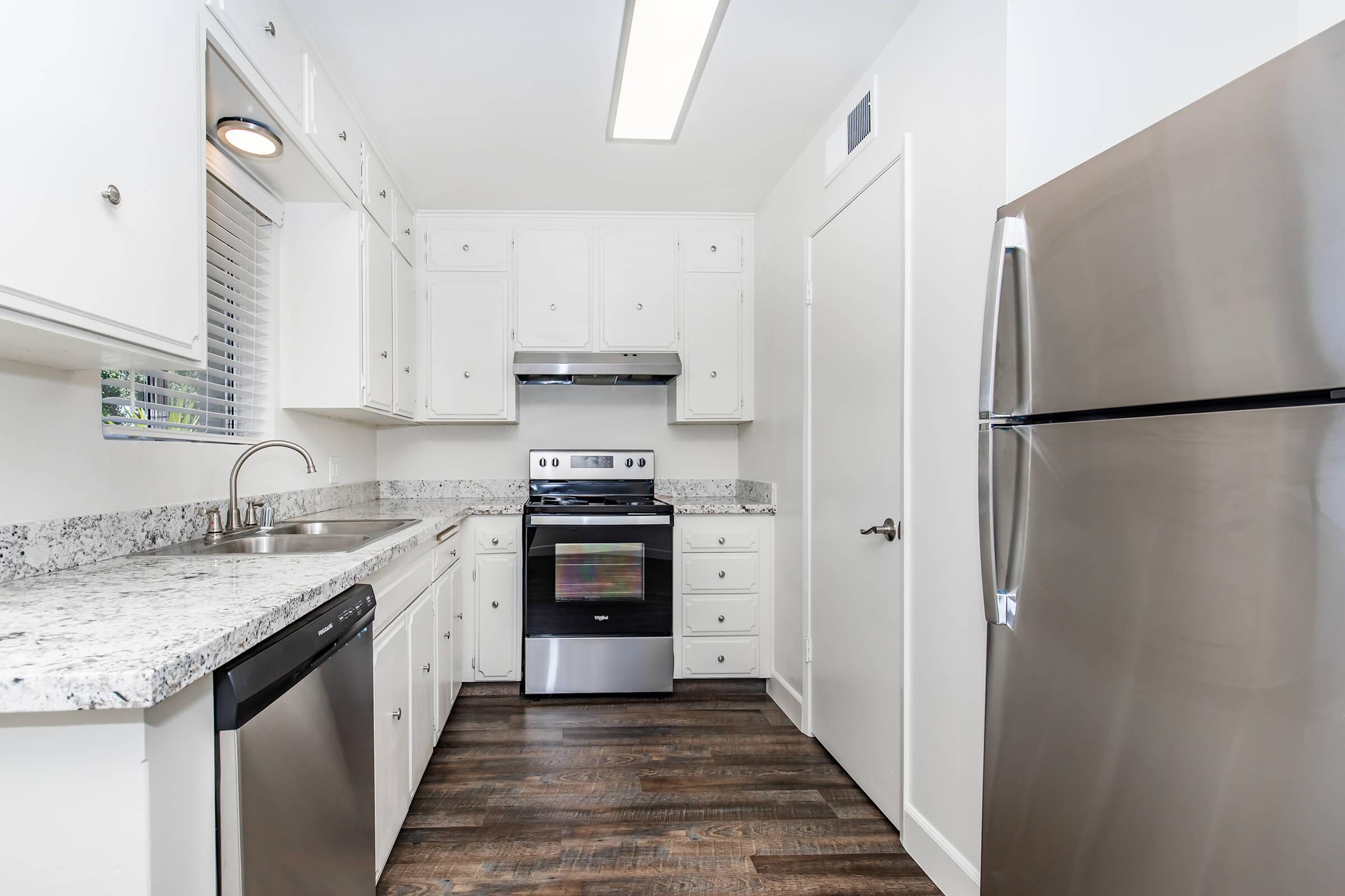 Modern kitchen featuring white cabinets, stainless steel appliances including a refrigerator and oven, granite countertops, and dark wood flooring. Natural light enters through a window, illuminating the space.