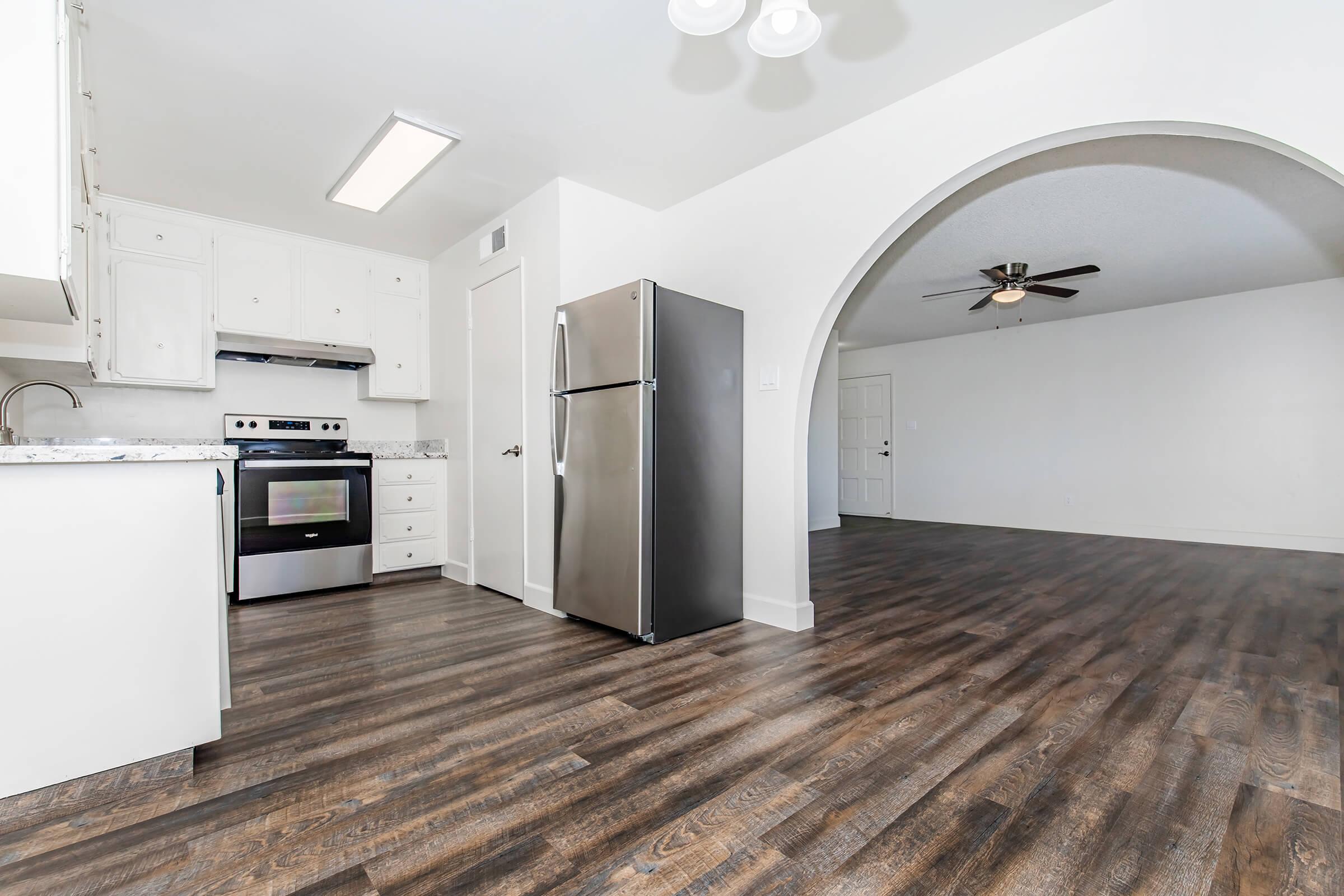 A modern kitchen featuring stainless steel appliances, including a refrigerator and oven, with white cabinetry and granite countertops. The open floor plan leads to a spacious living area with a ceiling fan. The flooring is dark wood, creating a warm contrast with the bright walls.