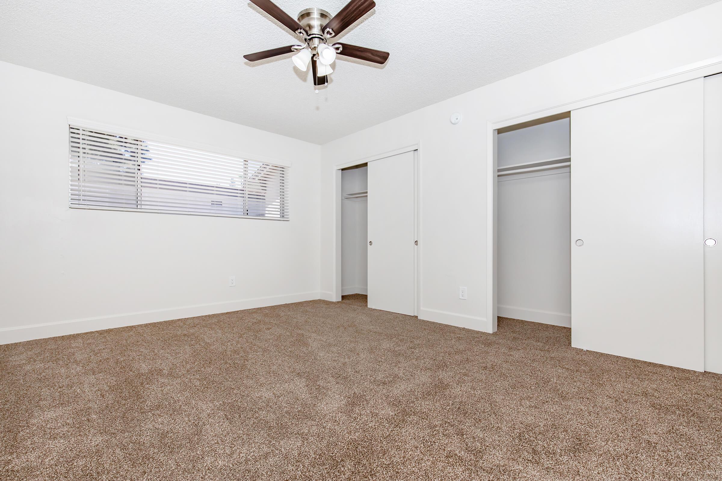 A clean, empty room featuring light-colored walls and a ceiling fan. There are two closed closet doors and a window with horizontal blinds, allowing natural light to enter. The floor is covered with soft, tan carpeting, enhancing the cozy atmosphere.