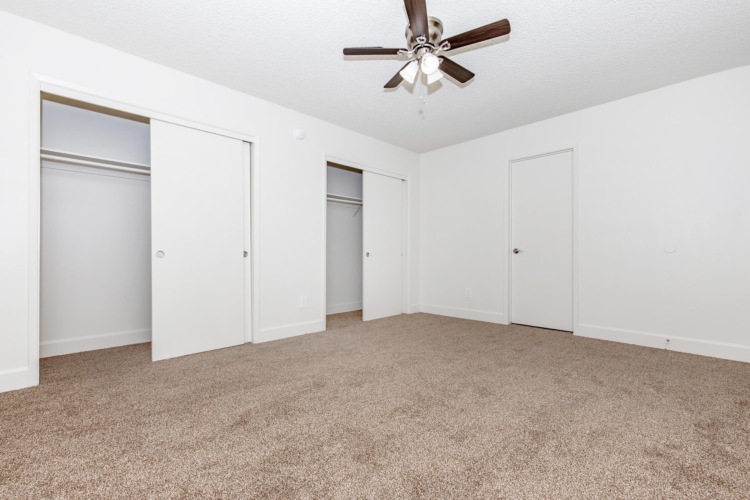 A spacious, empty room with light-colored walls and a textured ceiling. The floor is covered in beige carpet. There are two white closet doors on one side and a single door on the opposite side, along with a ceiling fan featuring dark wooden blades.