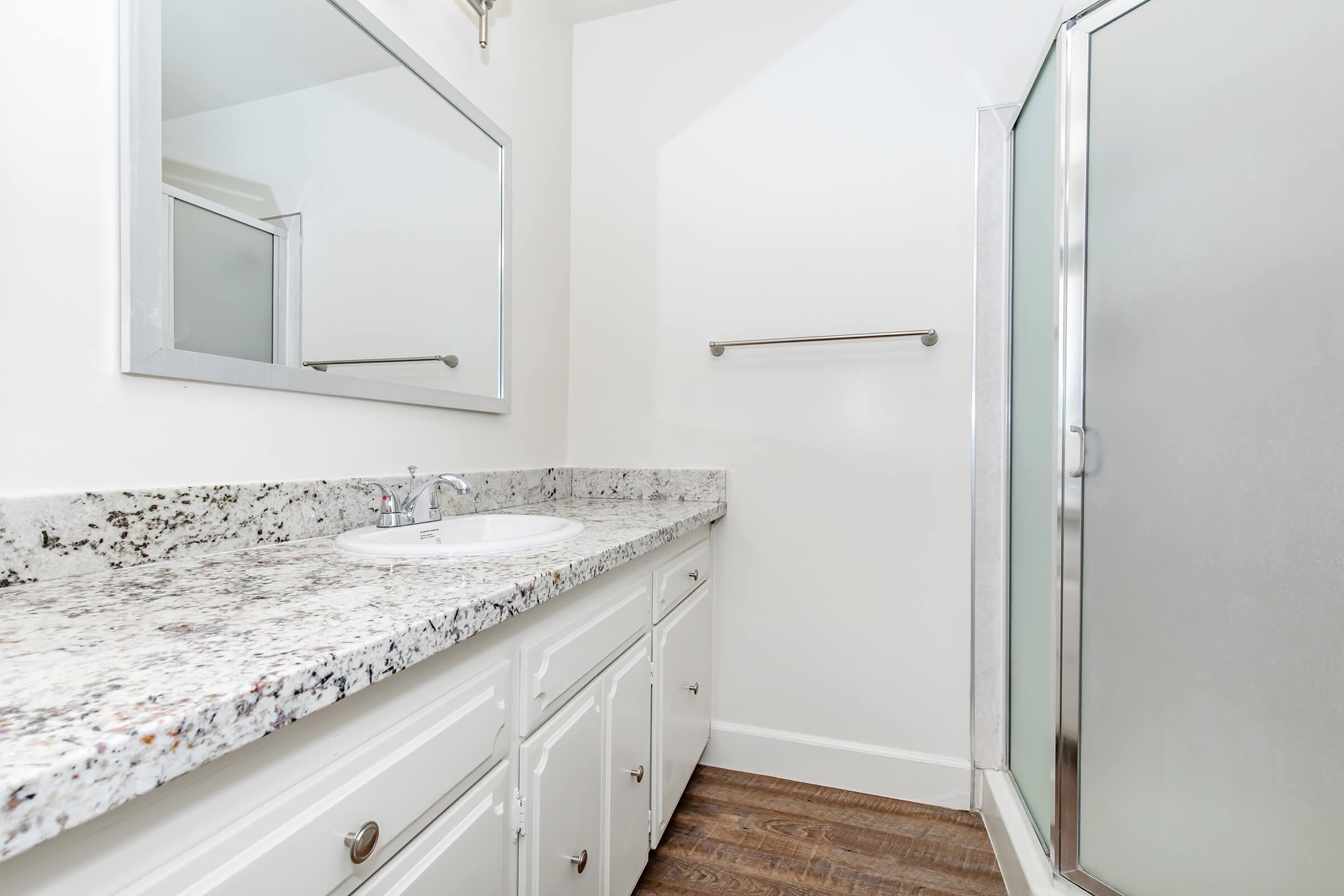 A modern bathroom featuring a granite countertop with a sink, white cabinetry, a large mirror above the sink, and a glass shower enclosure. The walls are painted white, and the floor has a wooden finish, creating a clean and contemporary aesthetic.