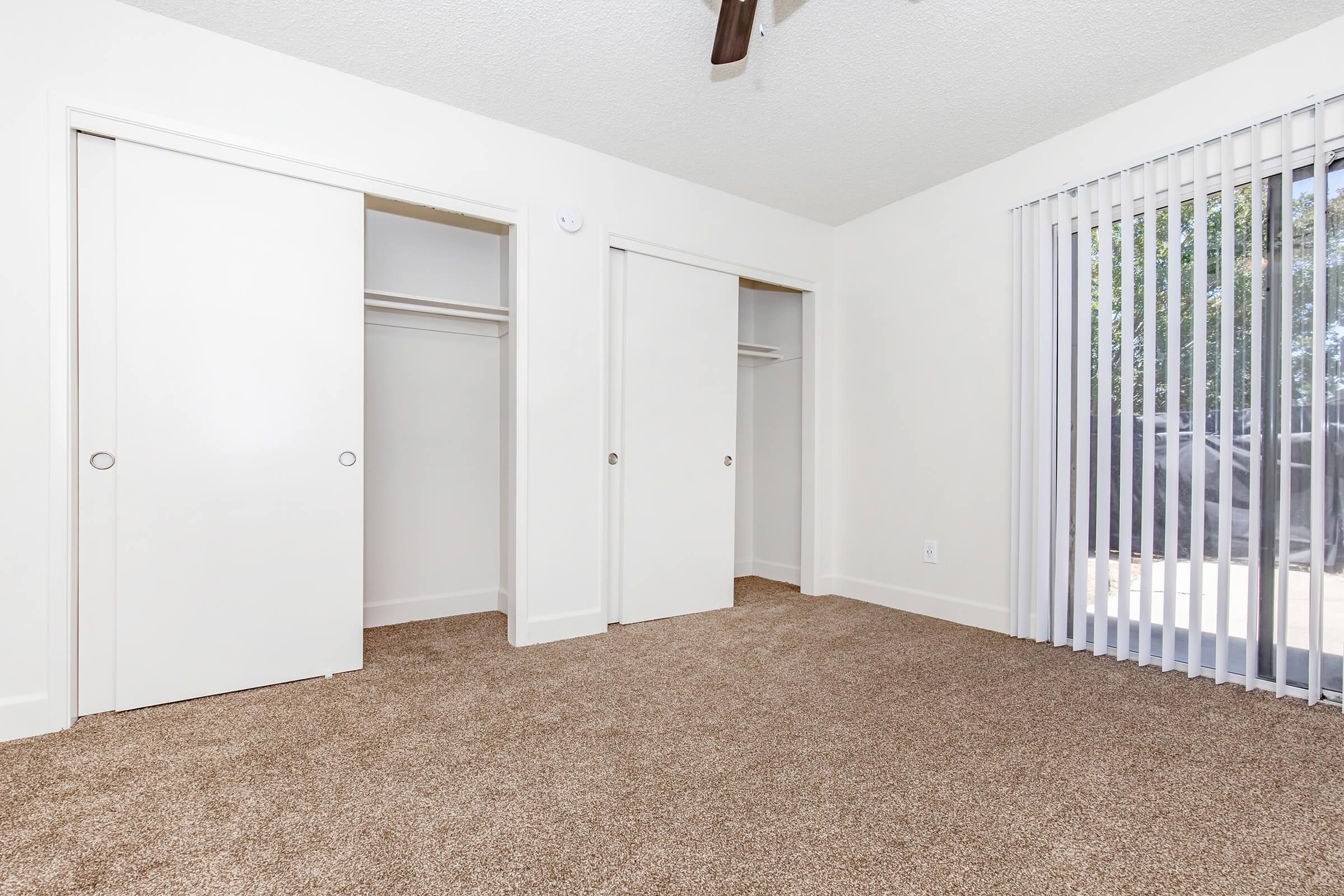 A spacious room featuring light-colored walls, two sliding closet doors on one side, and a large window with vertical blinds. The floor is covered with tan carpet, enhancing the room's bright and airy atmosphere. Natural light enters through the window, illuminating the space.