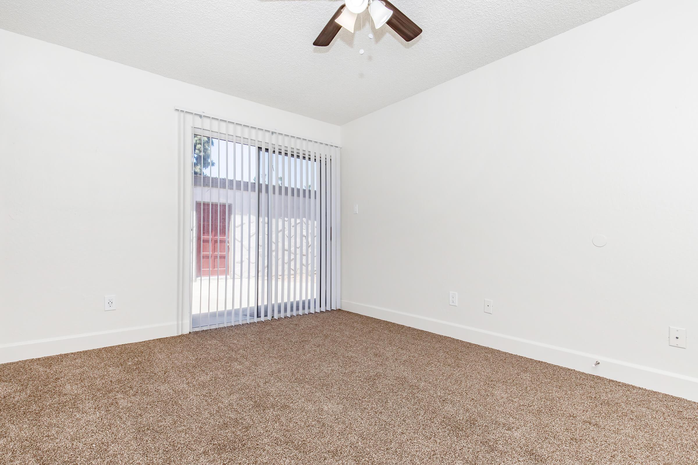 A bright, empty room with light beige carpet, white walls, and a ceiling fan. Large sliding glass doors with vertical blinds lead to an outdoor area, offering natural light. Minimalist design with no furniture present, creating an open and airy atmosphere.