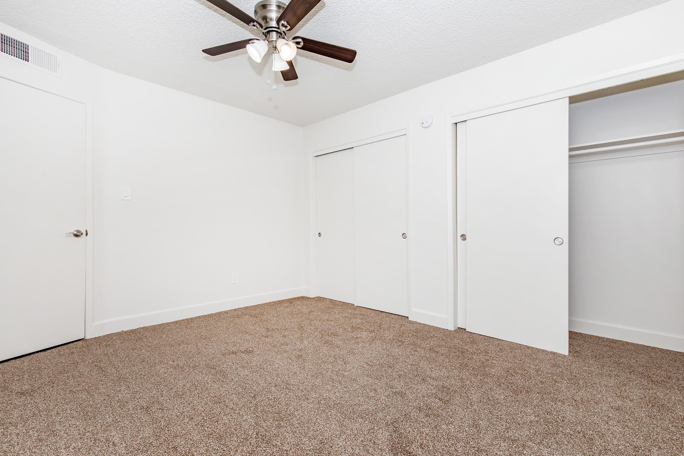 A well-lit, empty room with beige carpet flooring, featuring a ceiling fan and white walls. Two closed closet doors are visible on the right, and a door to the left leads to another area. The room has a clean, minimalist design with no furniture present.