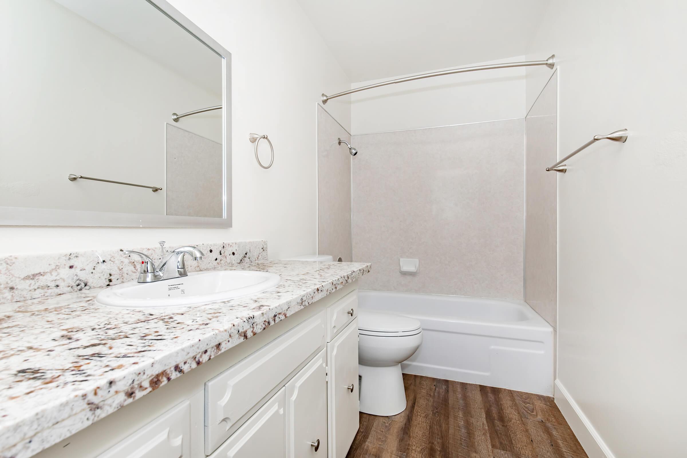 A clean and modern bathroom featuring a white bathtub and toilet, a granite countertop with a sink, a mirror, and light-colored walls. The space has a shower curtain and metallic towel bars, with a wood-like floor providing a warm contrast.